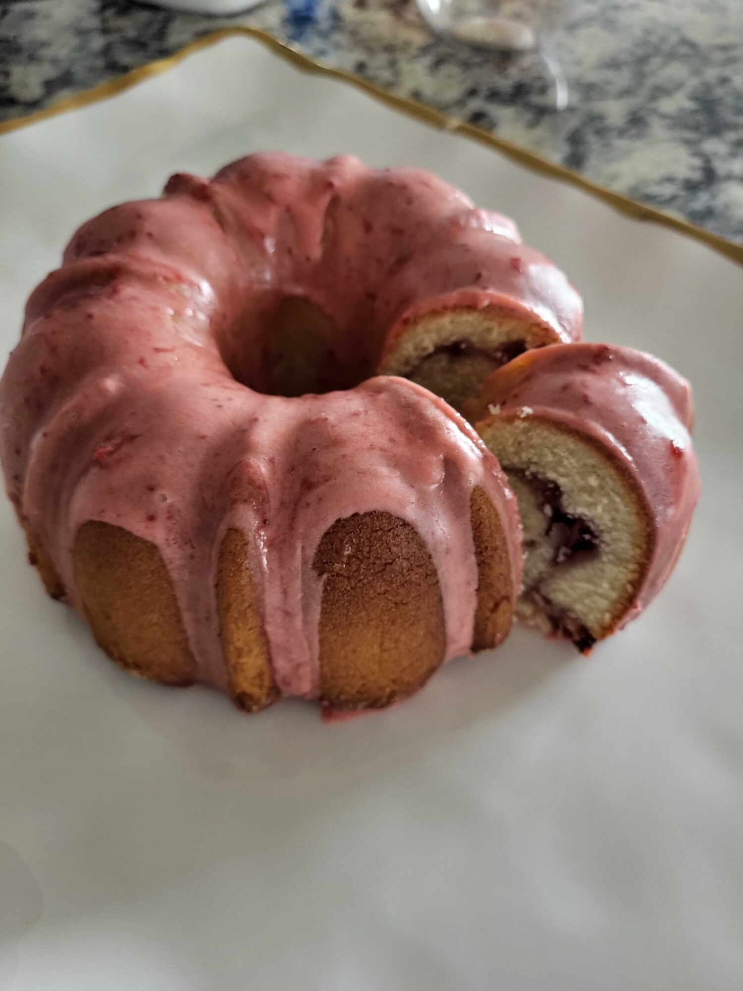 Pink glazed bundt cake with a slice showing berry filling, on a white plate with a gold rim, marble surface background.