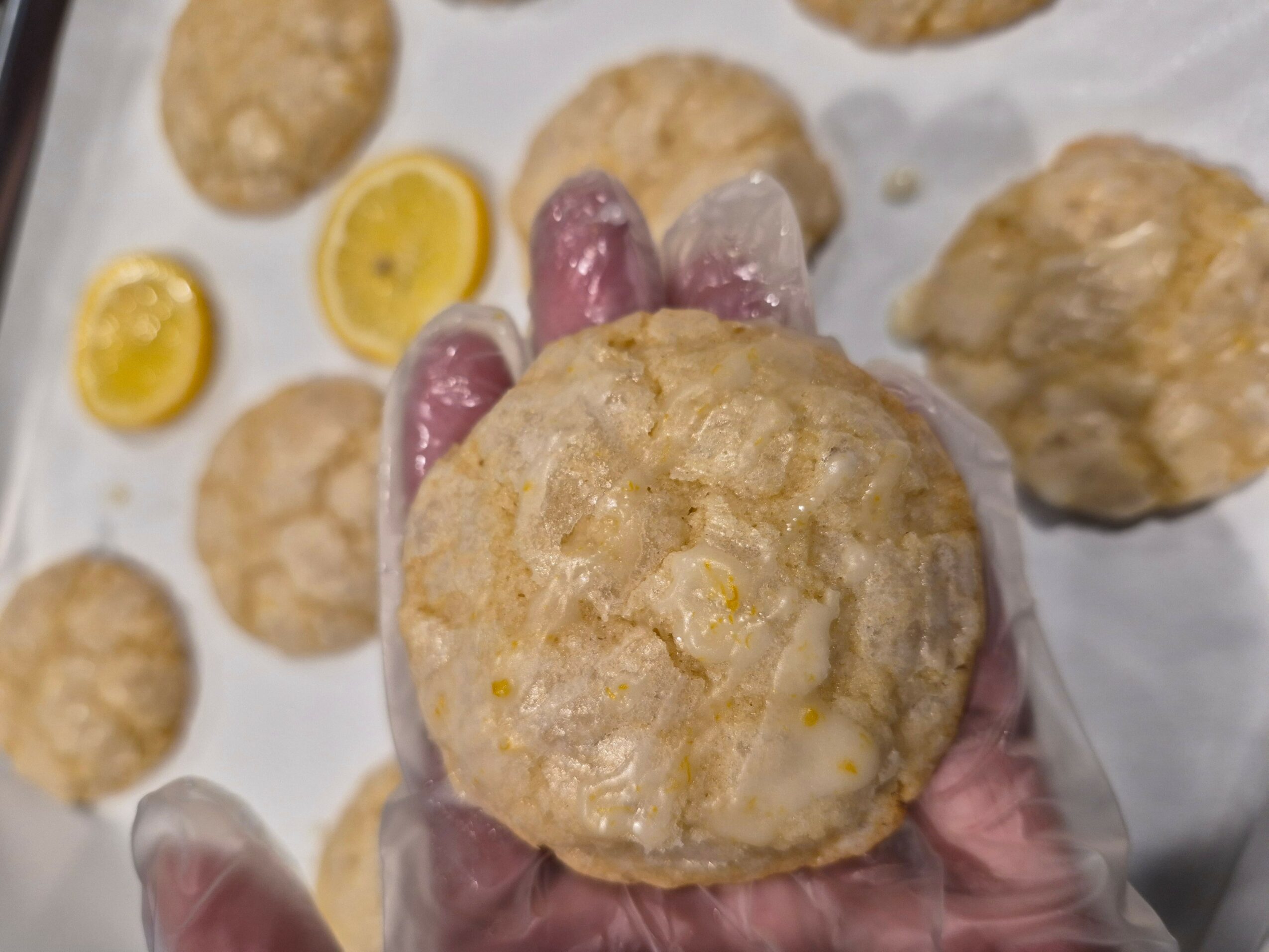 Hand holding a round, baked snack with a textured surface, surrounded by similar snacks and small yellow items on paper.