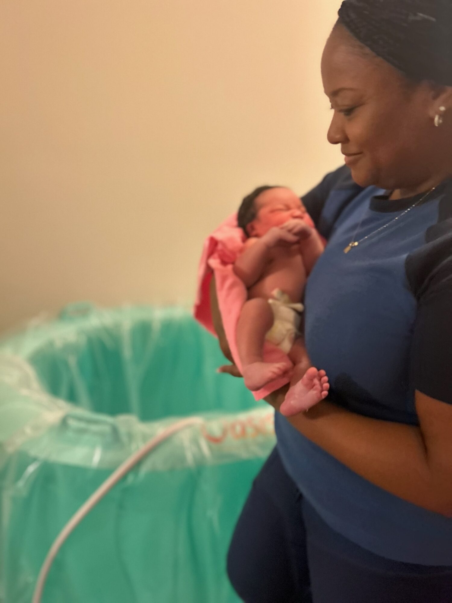 Woman holding a newborn baby near a hospital bassinet, smiling gently, in a clinical setting.