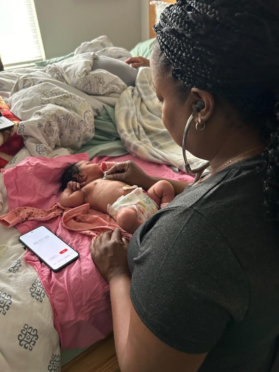 Woman with braided hair and earrings looks at a newborn baby on a bed, with a phone nearby, in a room with natural light.