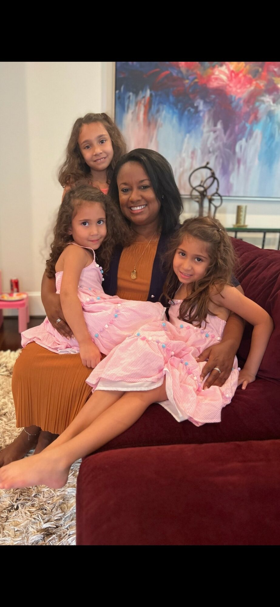 Four women sitting on a sofa, two children in pink dresses, smiling, in a living room with colorful artwork.
