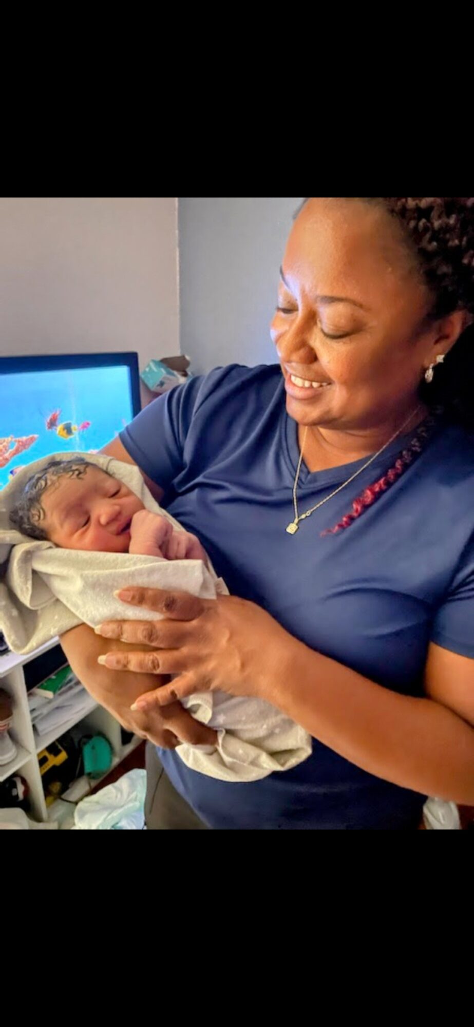A woman holding a newborn baby wrapped in a blanket, smiling warmly, in a room with a computer monitor in the background.
