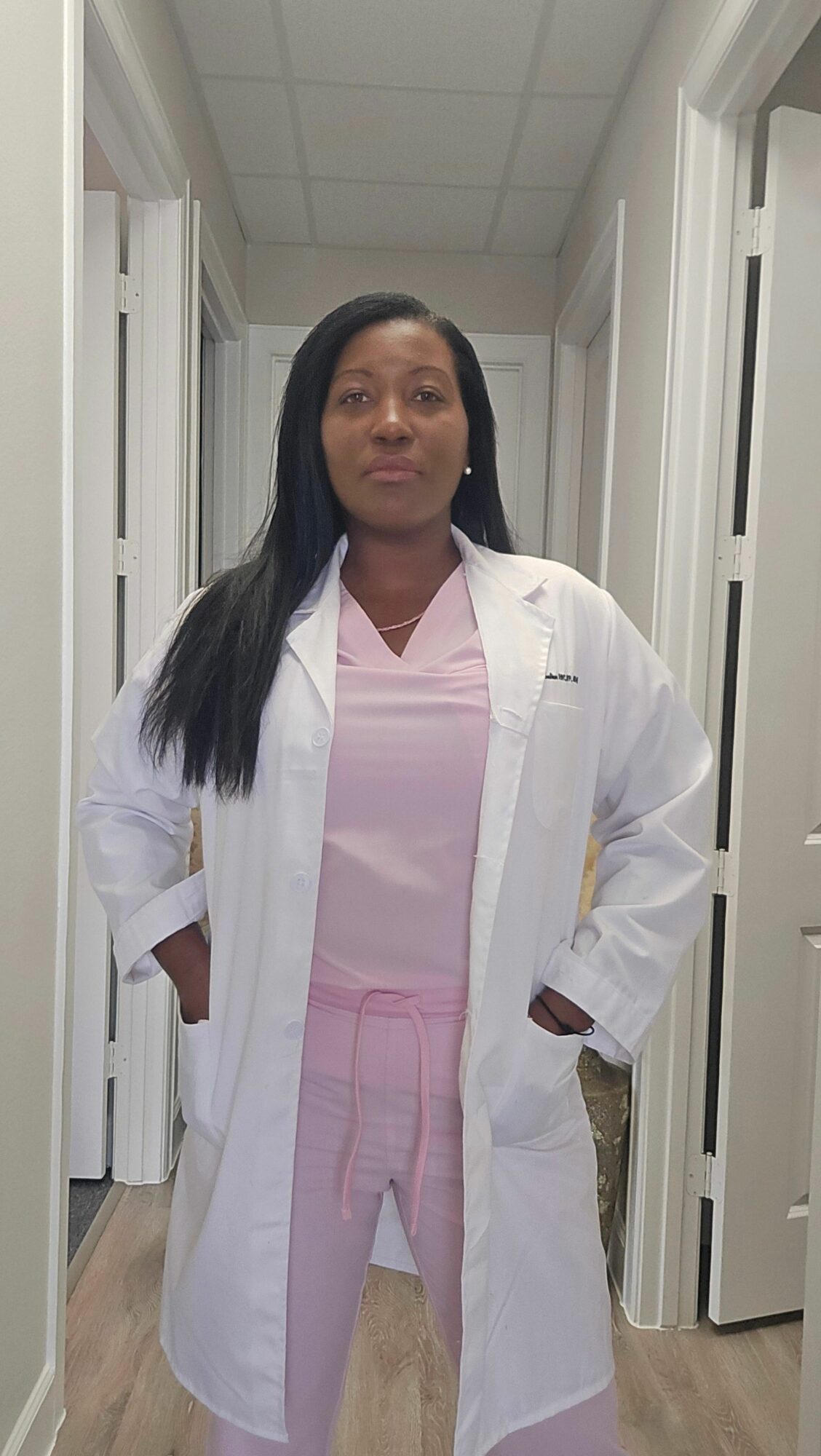Woman in white coat and pink dress standing indoors, facing camera, with long black hair, in a room with white walls and doors.