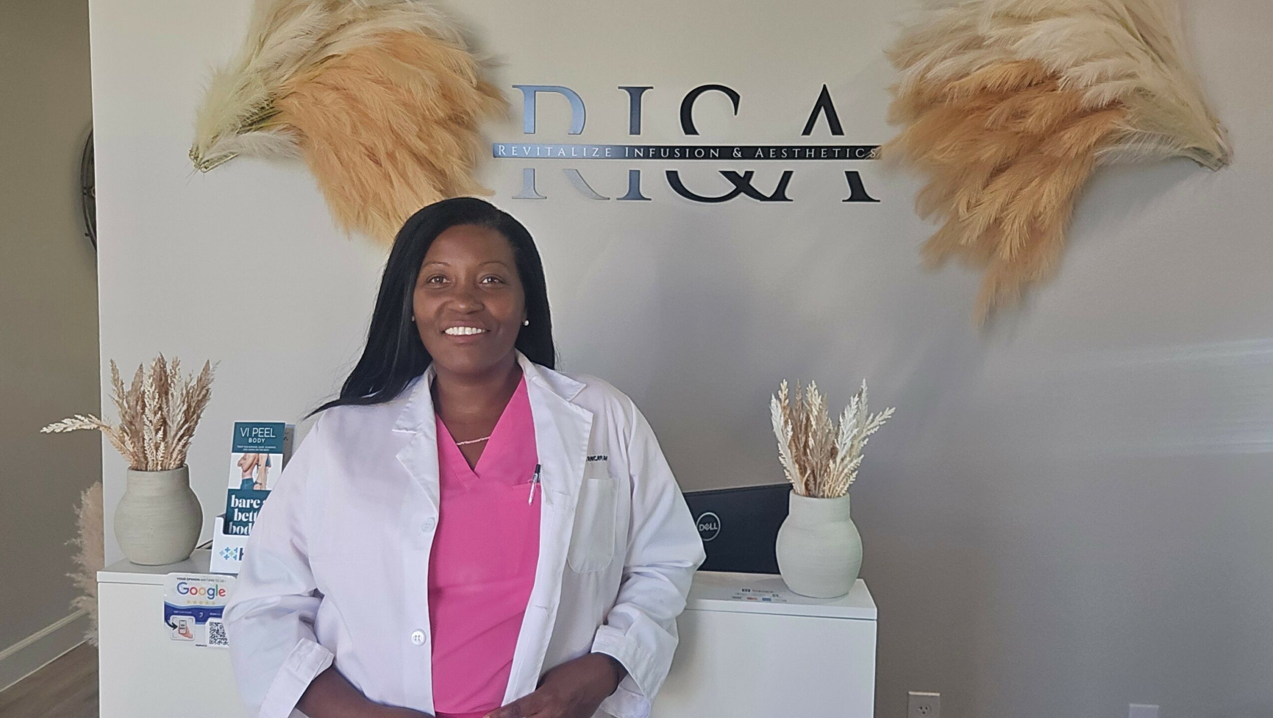 Woman in pink scrubs and white coat standing in front of a wall with decorative feathers and plants.