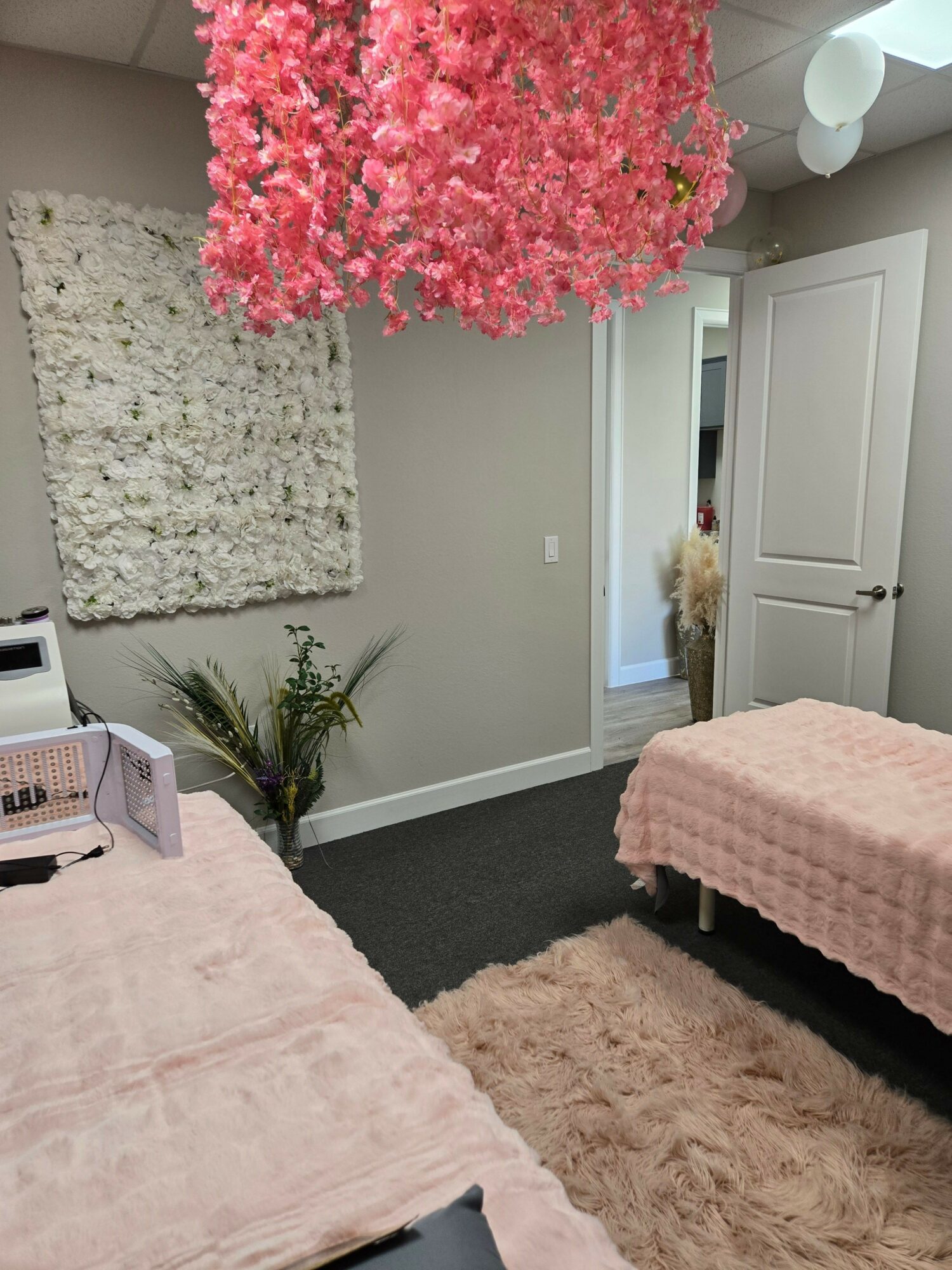 Corner of a bedroom with a pink bedspread, a beige rug, a potted plant, and a pink flowering tree near a door.