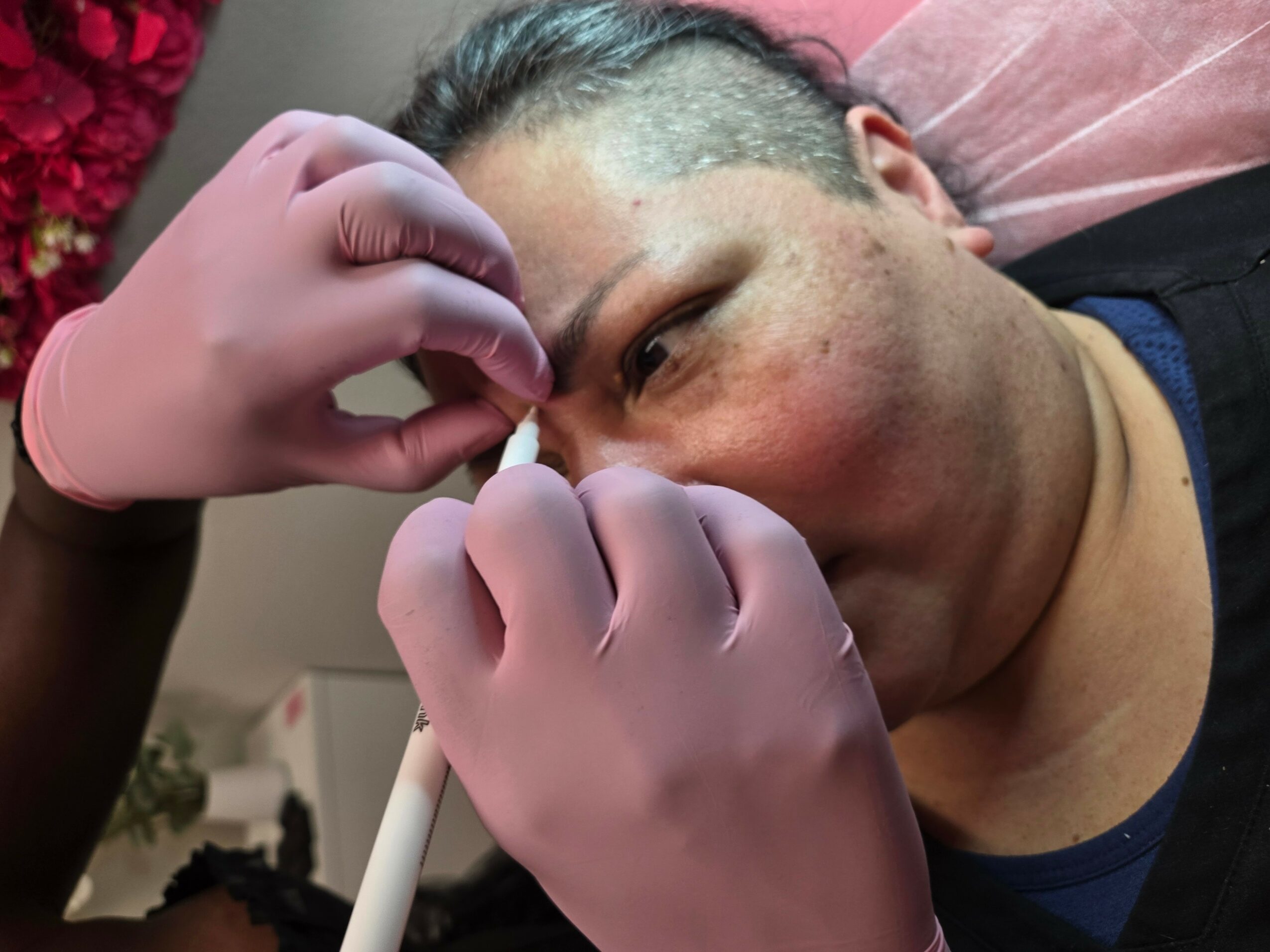 Person receiving a facial treatment with gloved hands near face, lying on a pink pillow, background with red flowers.