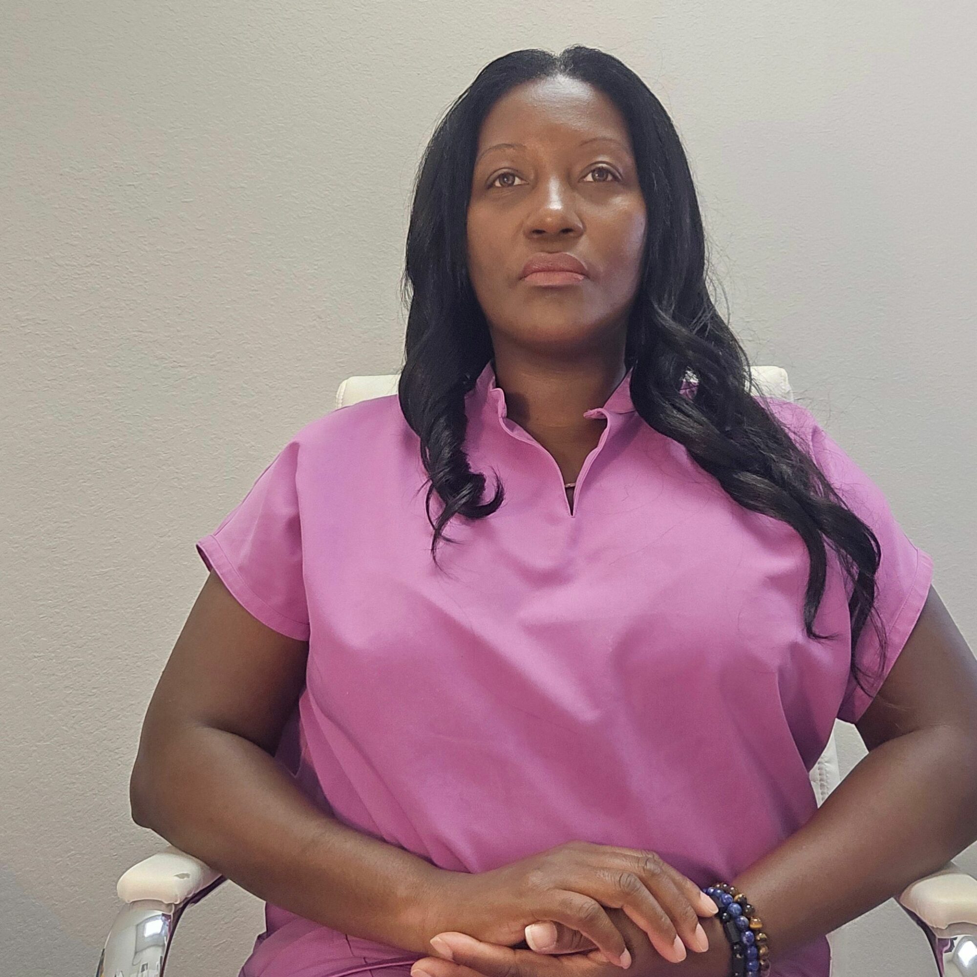 Woman with long black hair wearing pink shirt sitting against a plain wall, hands clasped, looking at camera.