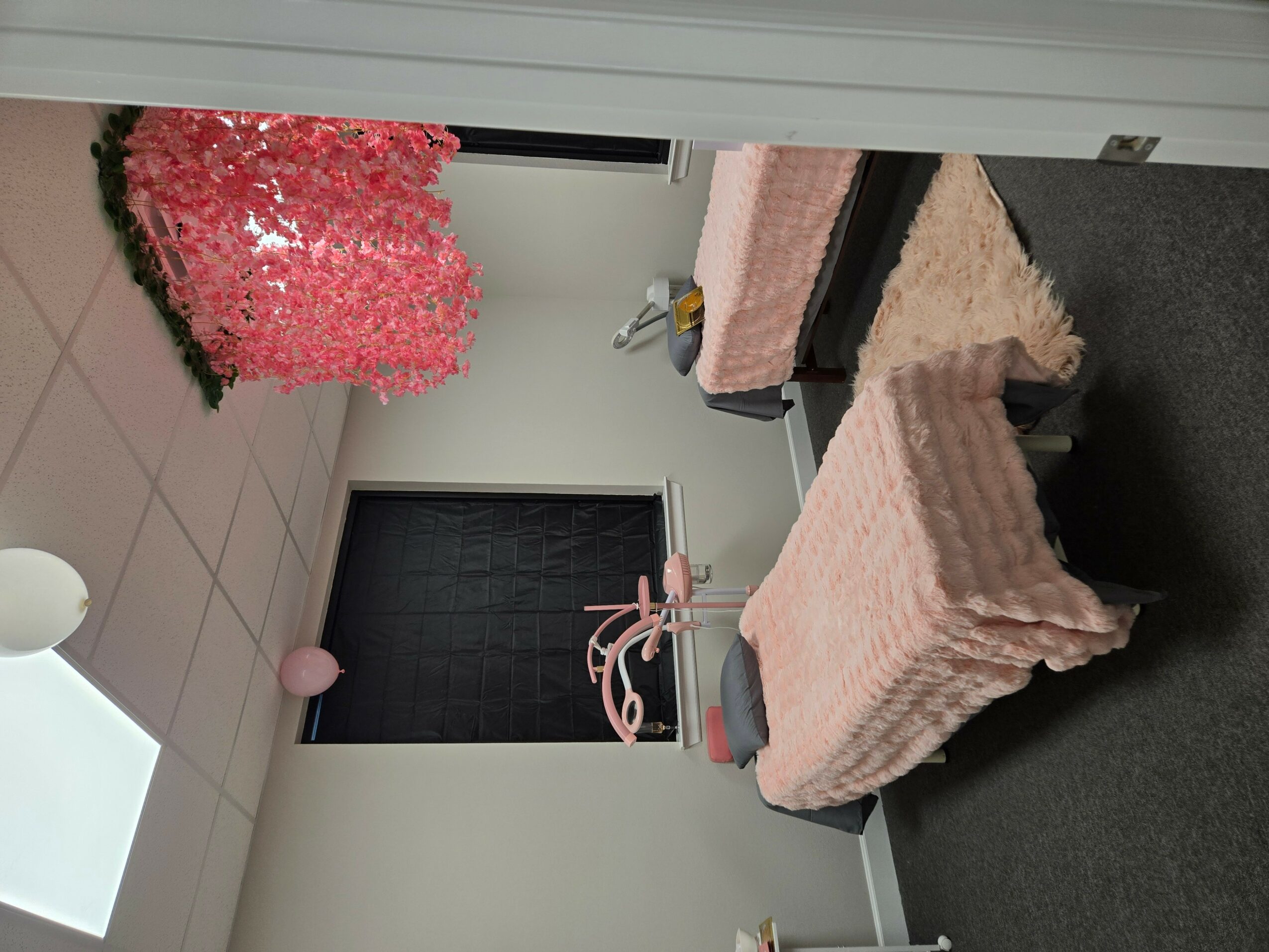 Room with a bed, pink blanket, pink lamp, pink tree, and a pink chair, with a black door and white walls.