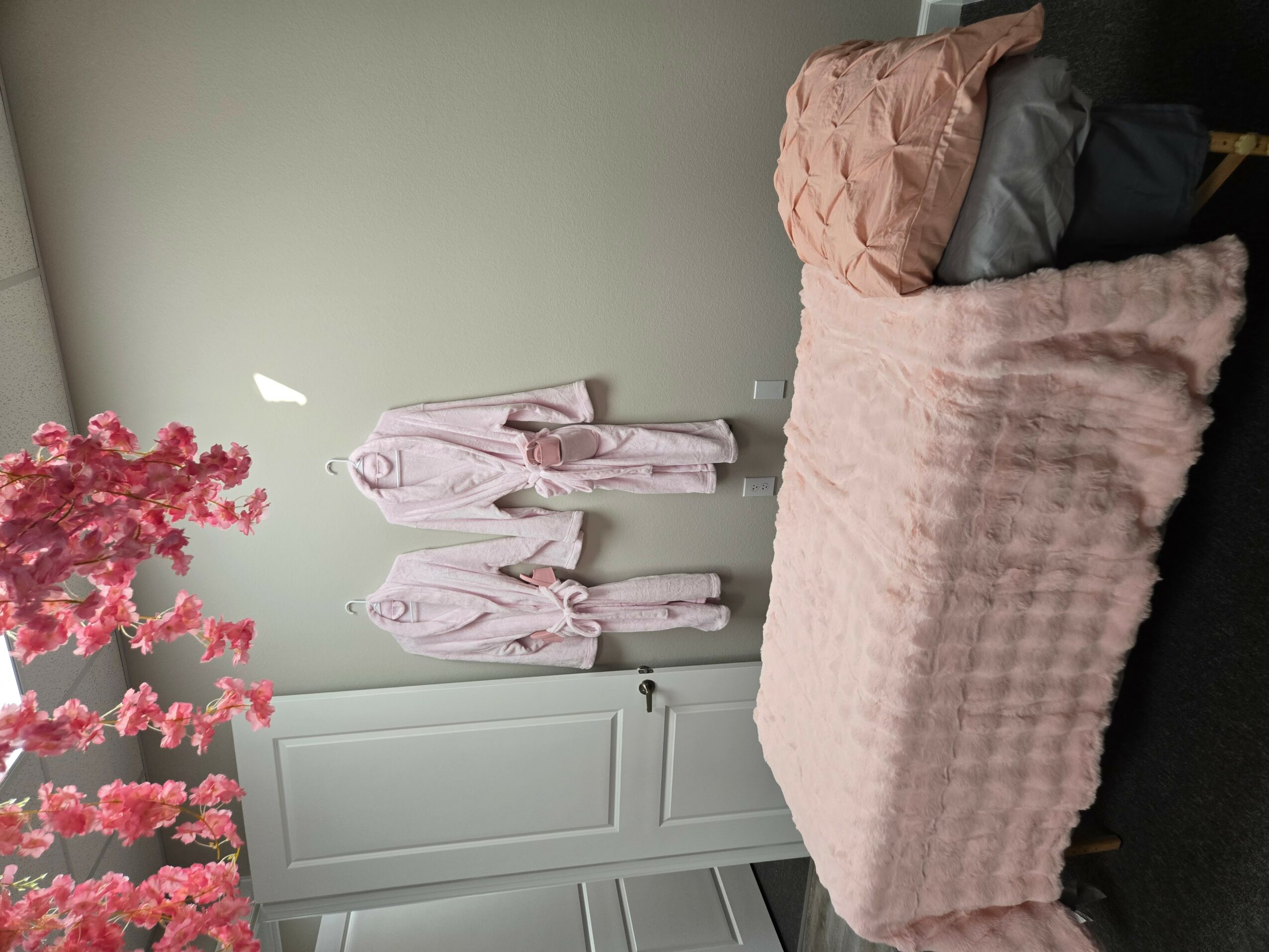 Bedroom with bed, pink blanket, pillows, and robes hanging on wall, pink flowers on left, light-colored wall, and dark floor.