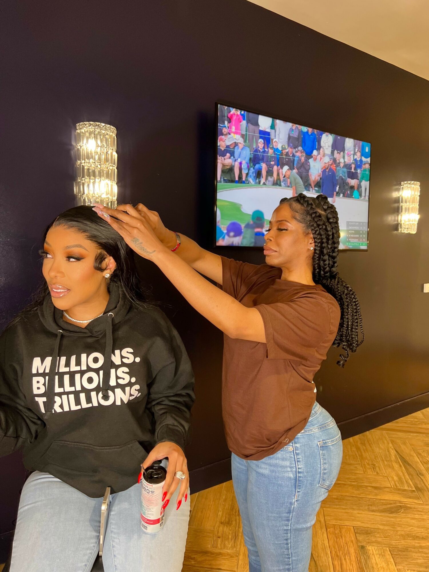 A woman styles another woman's hair in a room with a TV and wall lights, woman with long dark hair, other with curly hair.