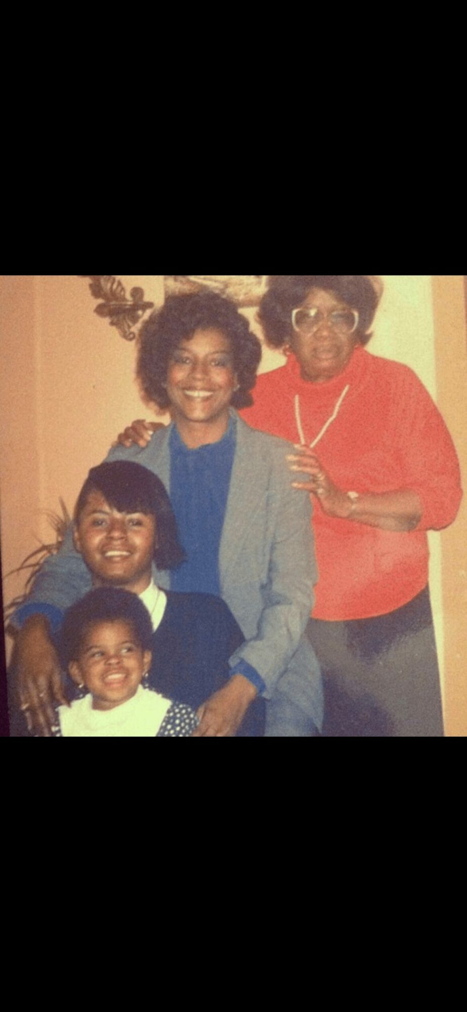 Four people smiling, two adults and two children, posing indoors with a decorative wall hanging behind them.