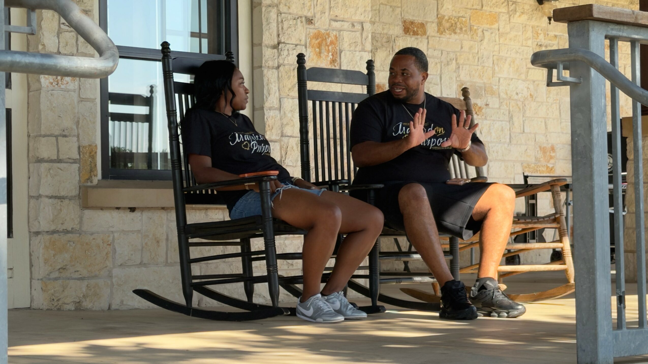 Two people sit on a bench outdoors, engaged in conversation, with a stone wall behind them.