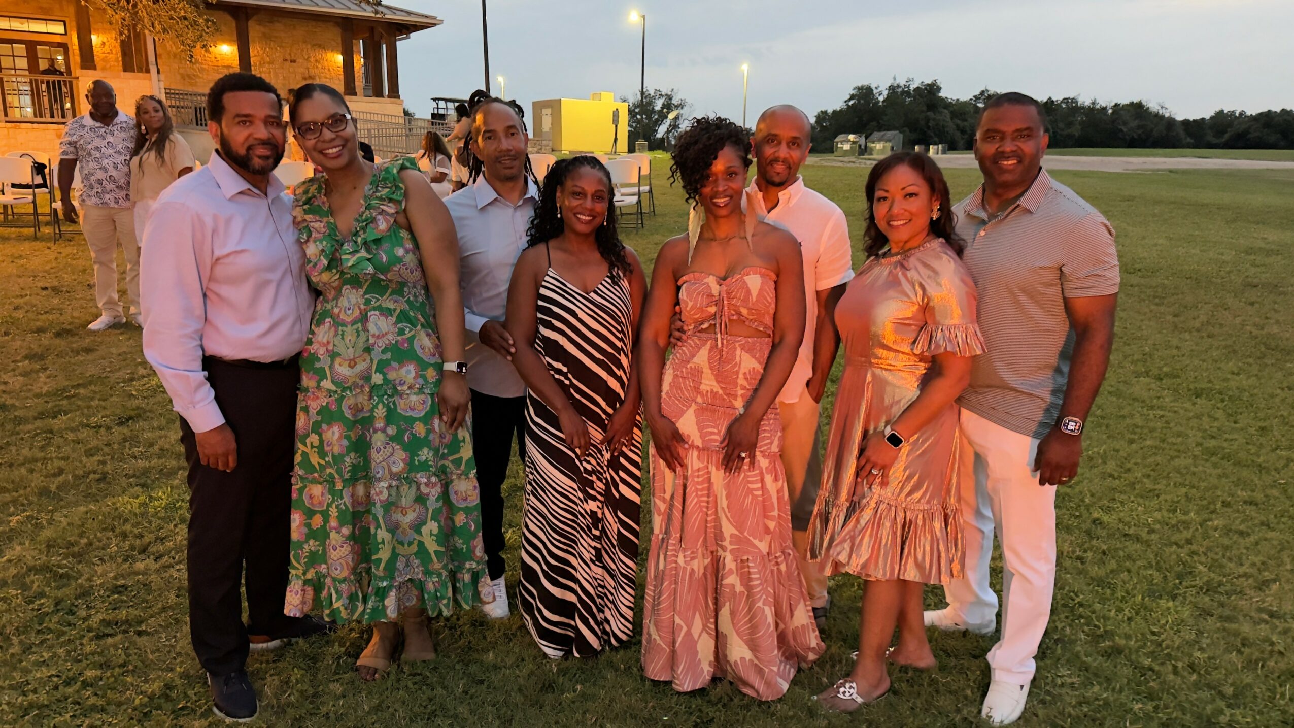 Group of nine people standing outdoors on grass, dressed in formal and semi-formal attire, smiling at the camera.