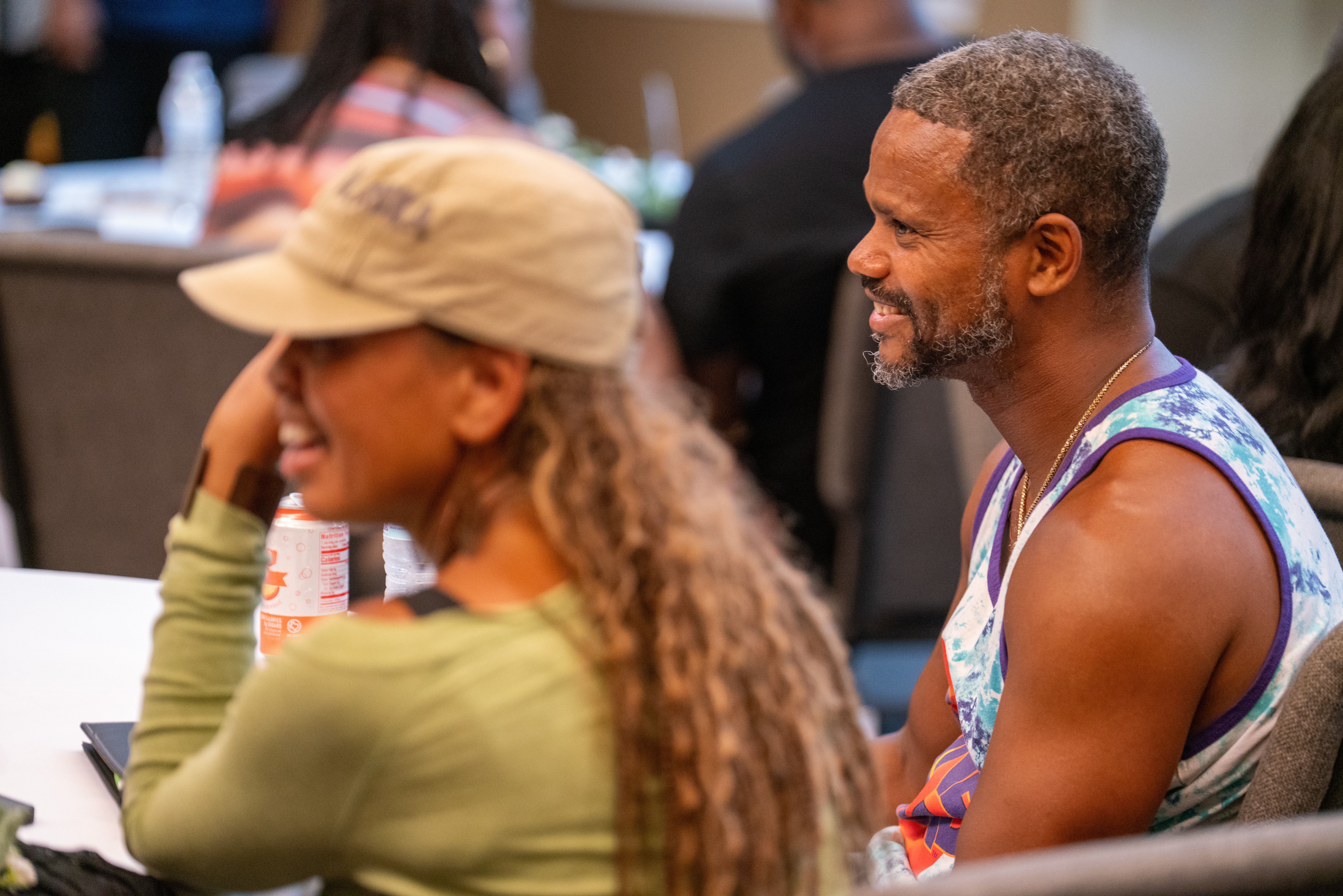 Two people sitting at a table, smiling and engaging in conversation, with others in the background.