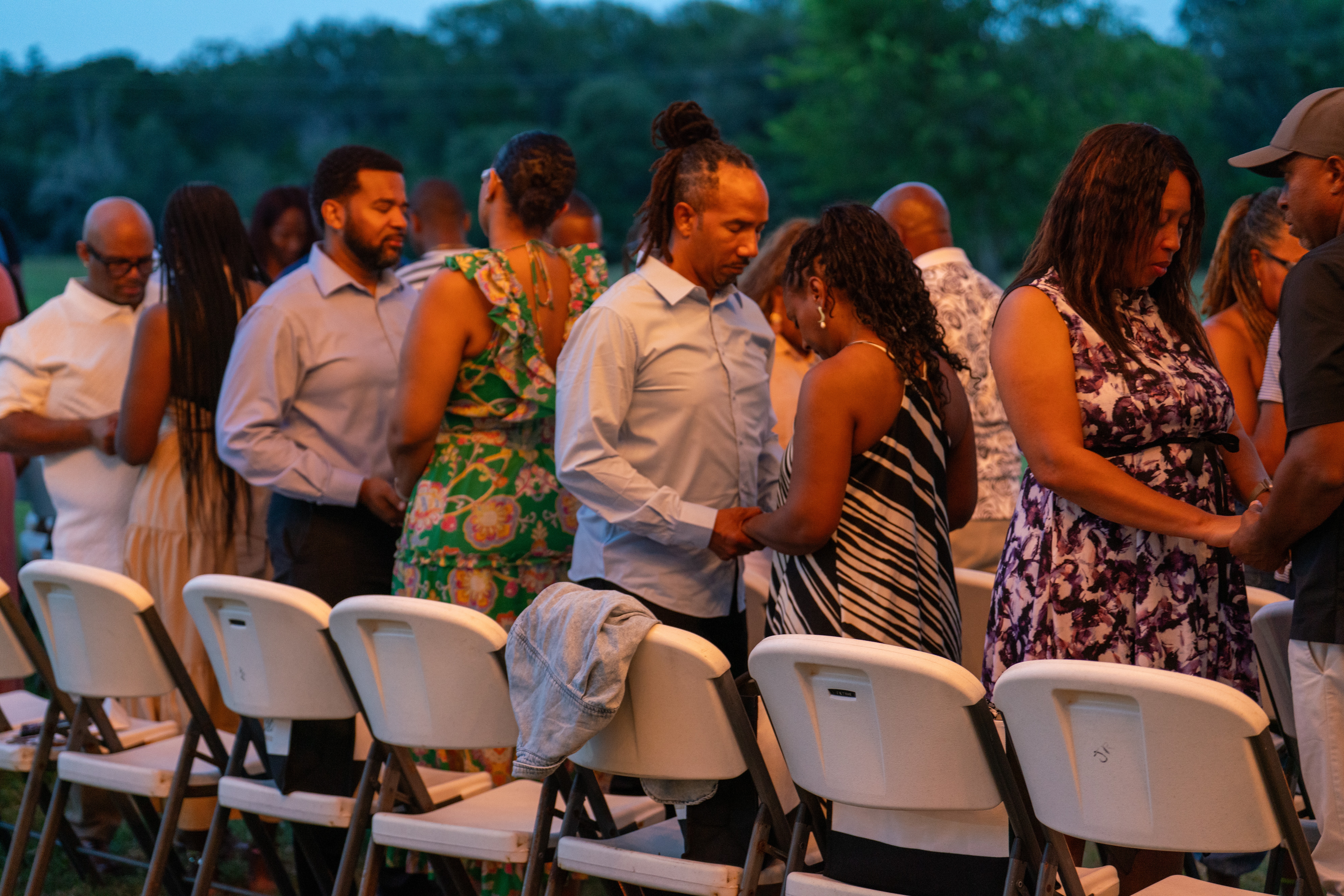 People standing in line outdoors, some holding hands, with chairs in front and trees in the background.