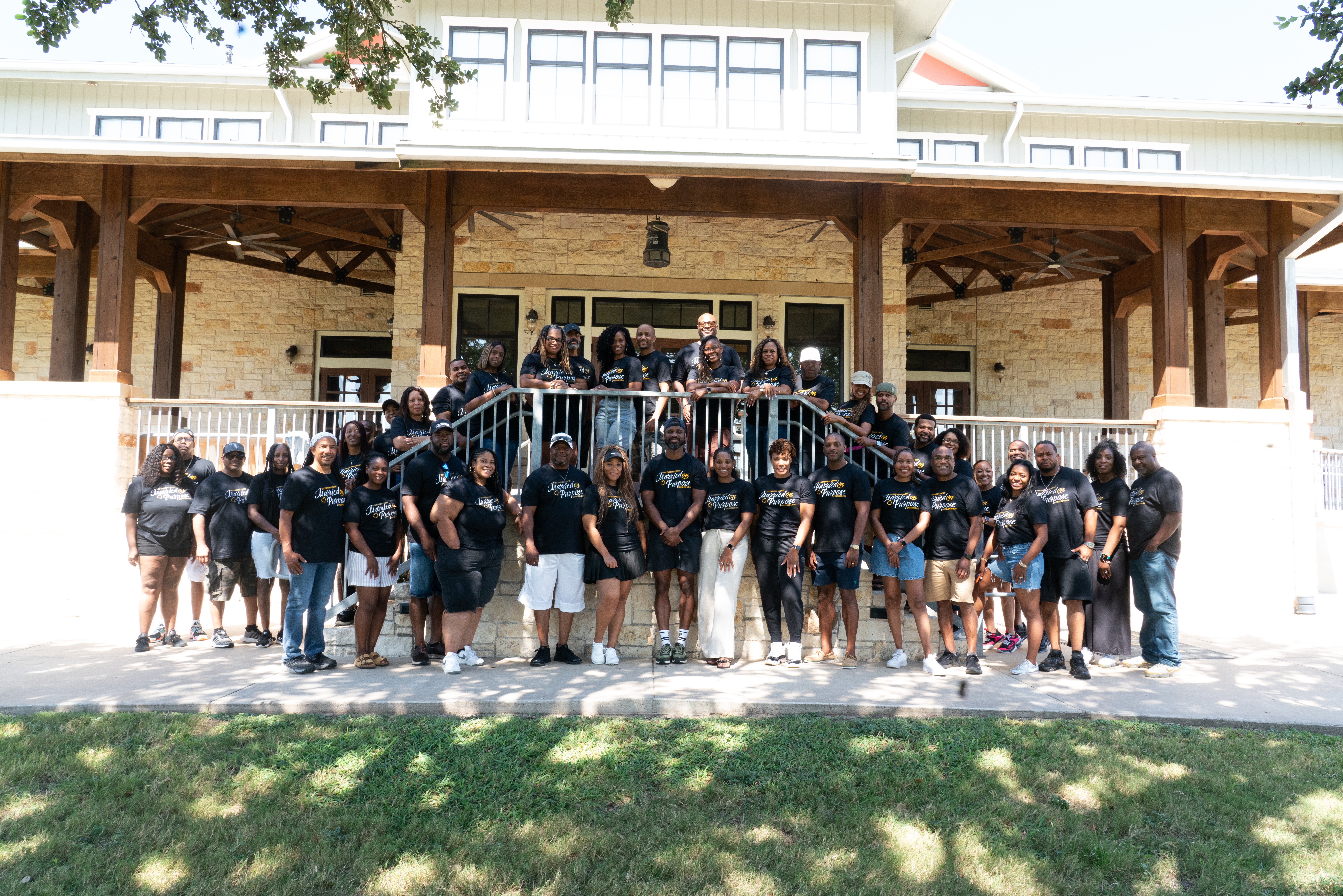 Group of people gathered outside a building with a wooden porch, some standing on stairs, others on the ground, in front of a grassy area.