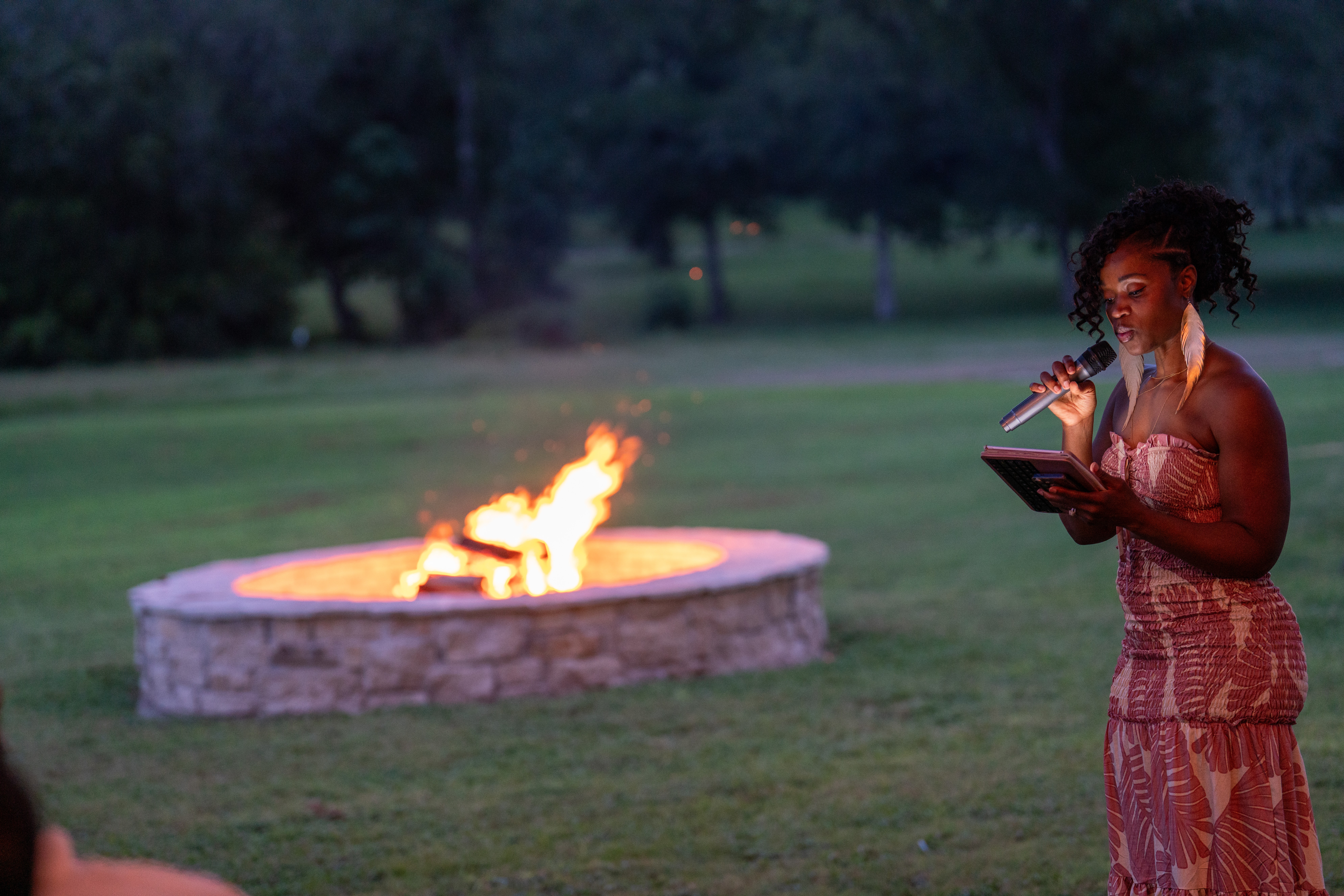 Woman reading a book near a fire pit outdoors at night, with trees in the background.