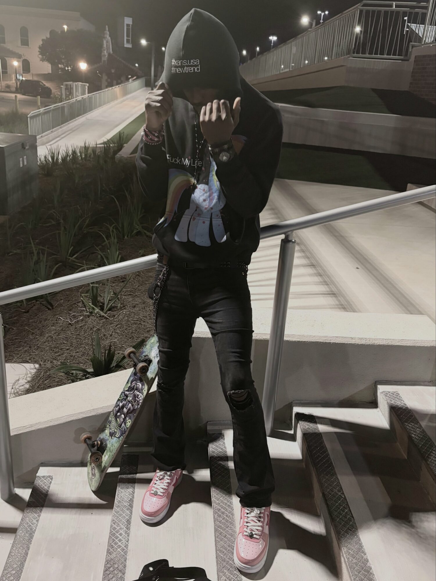 Person standing on stairs holding skateboard, face covered with hood, nighttime setting, urban environment.