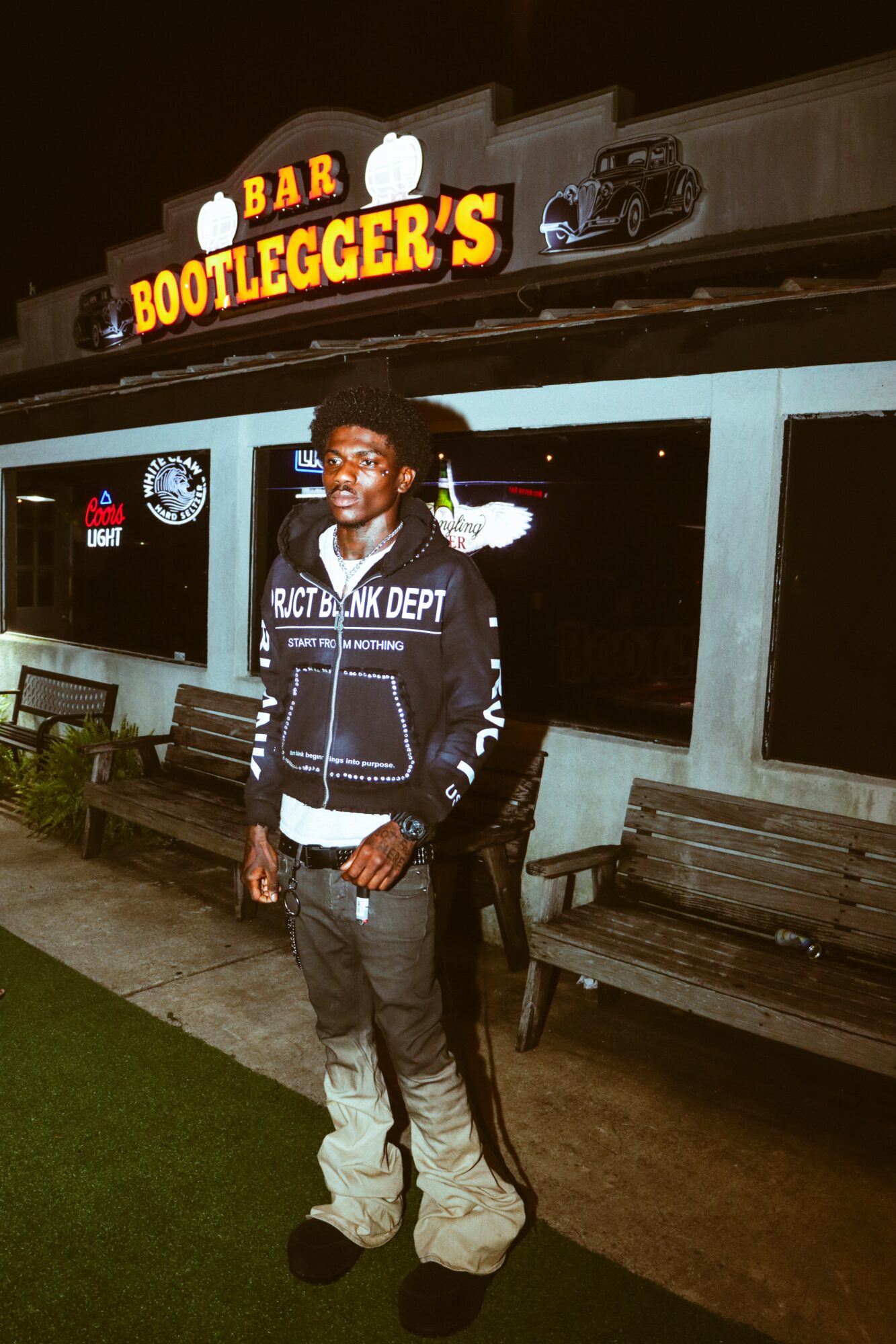 Young man standing outside a bar at night, illuminated sign above reads 'Bootlegger's' with a beer mug icon, benches nearby.