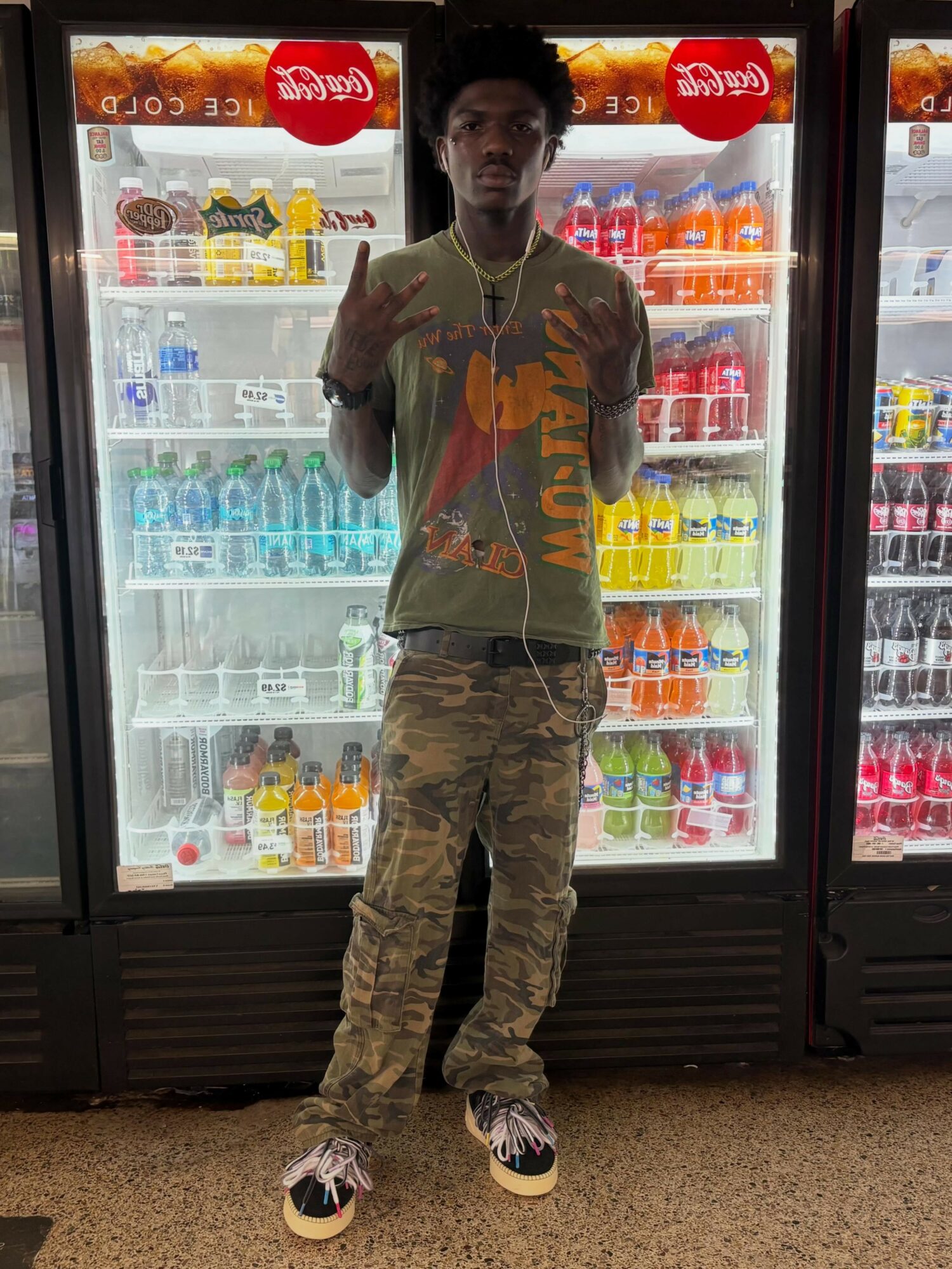 Young man standing in front of refrigerated beverage shelves, wearing camouflage pants and a colorful shirt, making hand gestures.