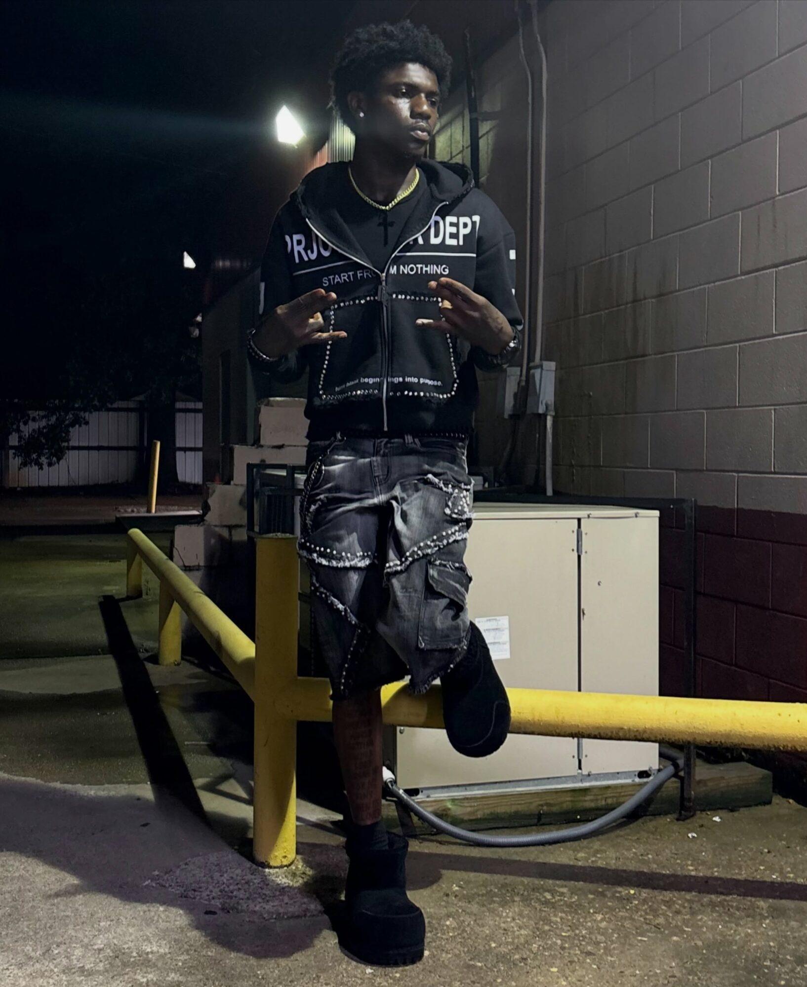 Person taking a mirror selfie outdoors at night, wearing a black jacket, shorts, and boots, holding a phone, with a yellow barrier nearby.