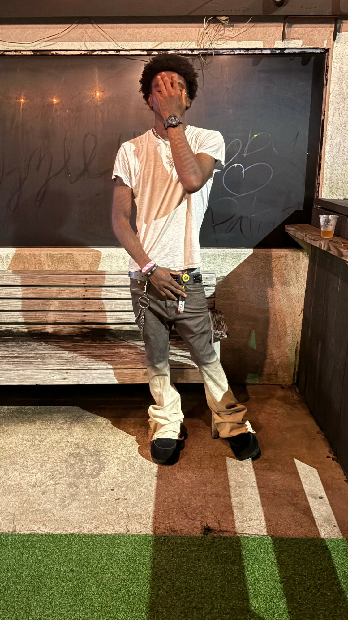 Young man with curly hair in a white t-shirt and baggy pants standing outdoors, covering his face with his hand.