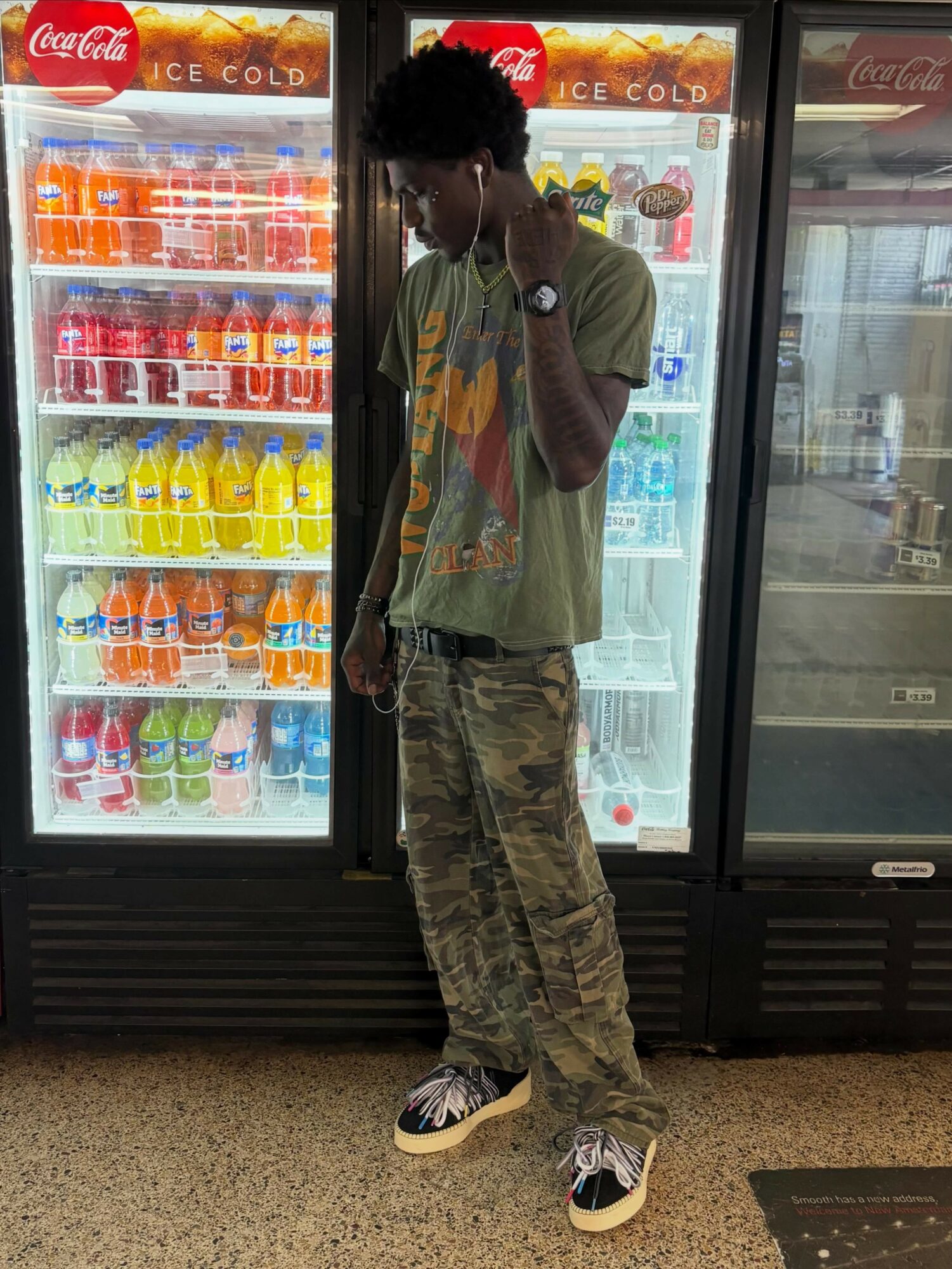 Young person with curly hair wearing a green t-shirt, camouflage pants, and sneakers, standing in front of beverage refrigerators.