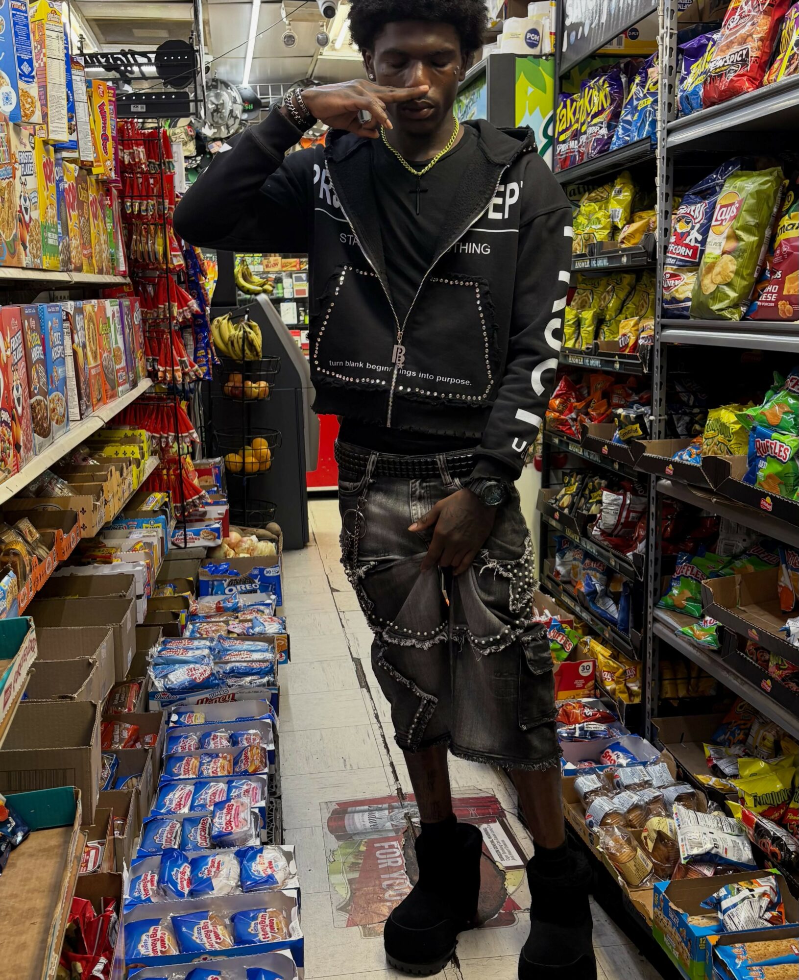 Young man with curly hair shopping in a store aisle filled with snacks and candies, wearing dark clothing and chains.