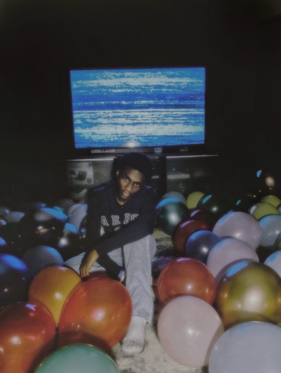 Person with glasses and dark shirt standing among colorful balloons in front of a large screen, in a dark room.