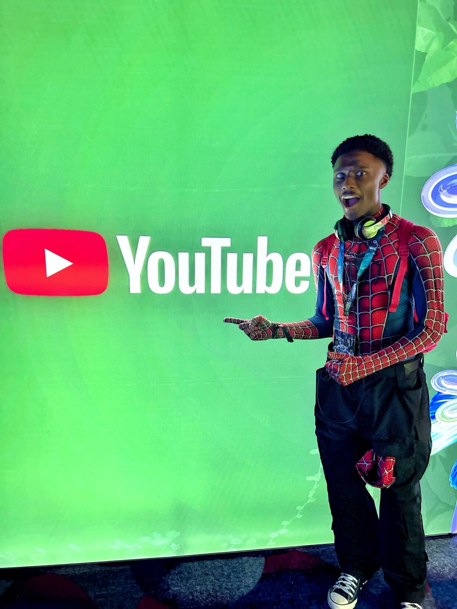 Young person standing in front of a green background with YouTube logo, wearing headphones and casual clothing.