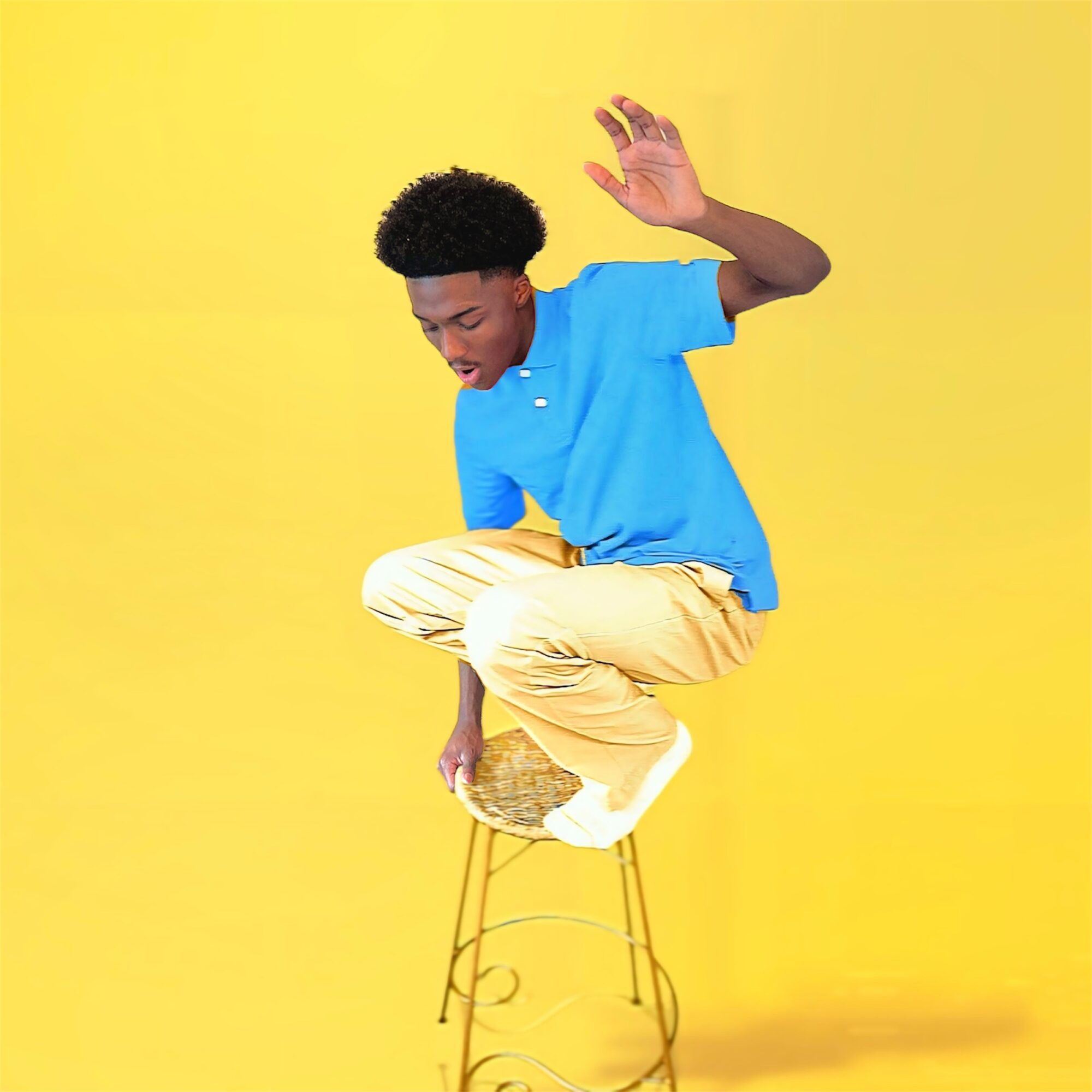 Young person balancing on a stool with a yellow background, wearing a blue shirt and beige pants, raising one hand.