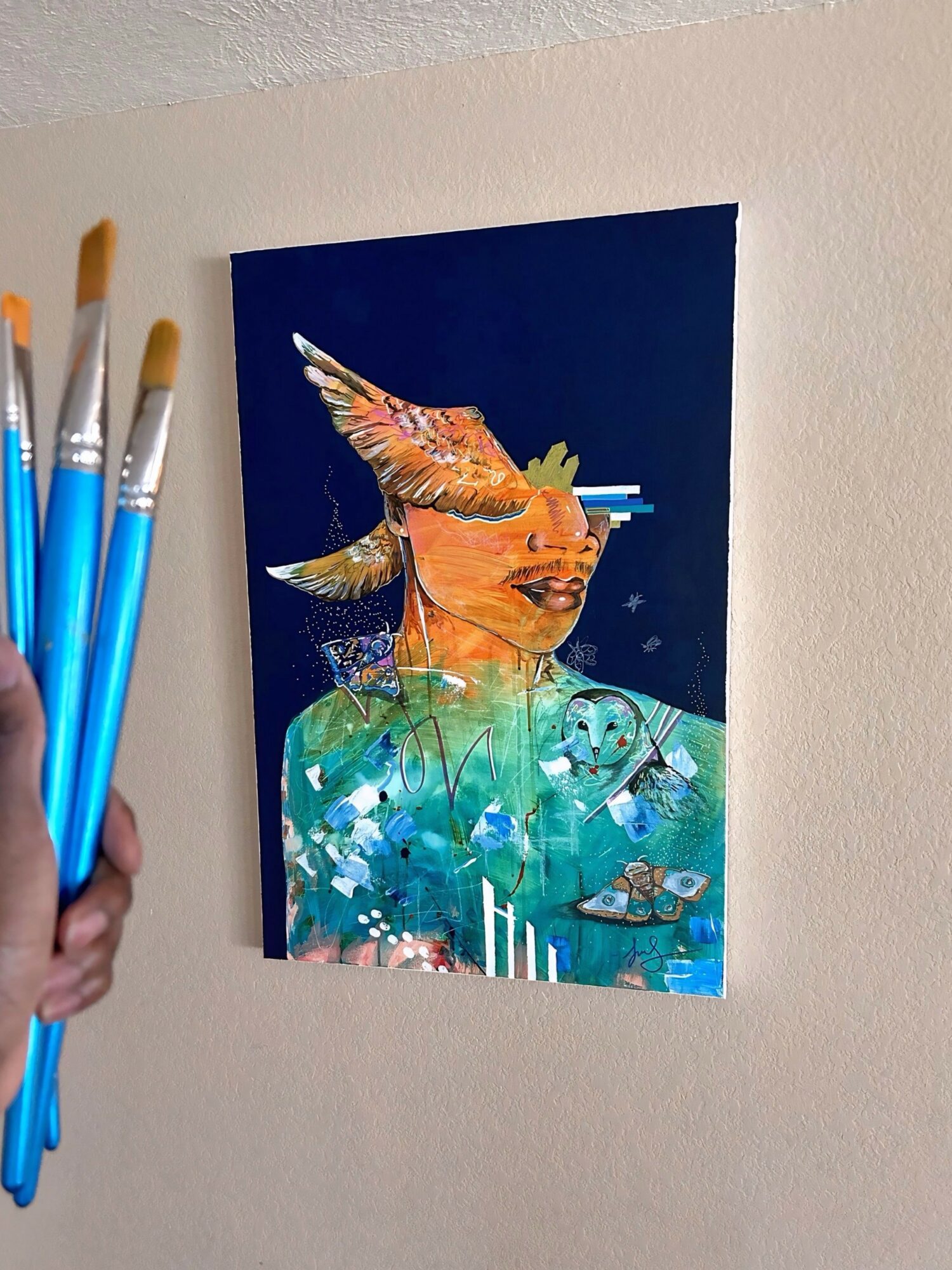 Colorful mixed media artwork of a person's face with bird wings, set against a dark background, held by a hand with paintbrushes.