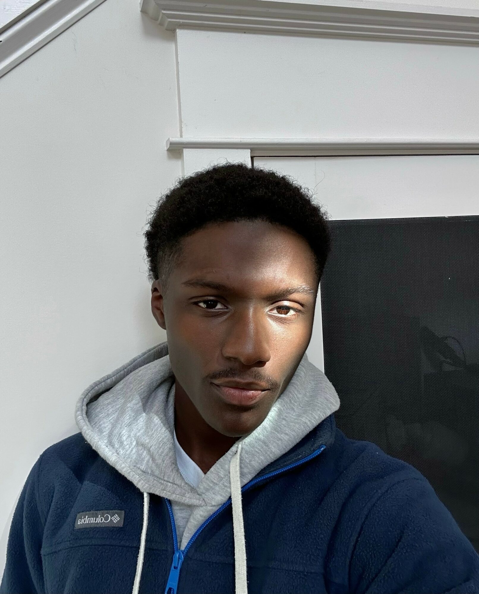 Young man with short curly hair wearing a hoodie and jacket, standing indoors near a black screen or monitor.