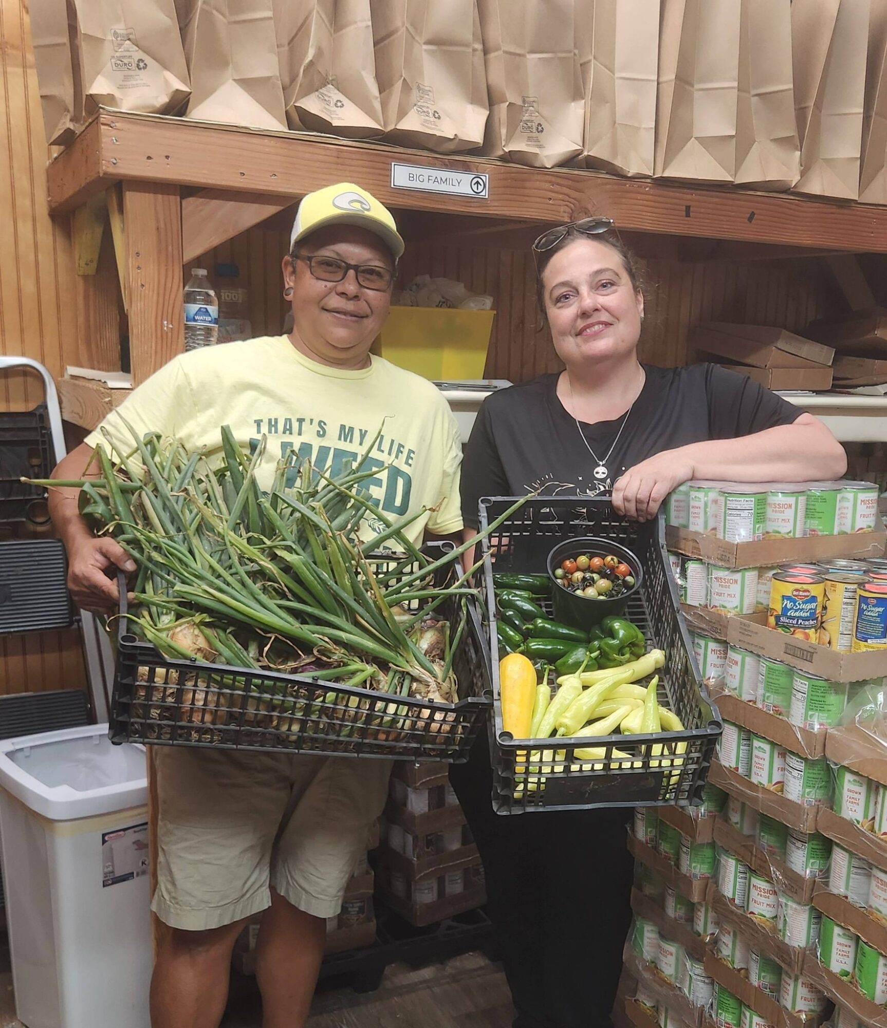 Two people standing in a storage area holding baskets of vegetables, smiling at the camera.