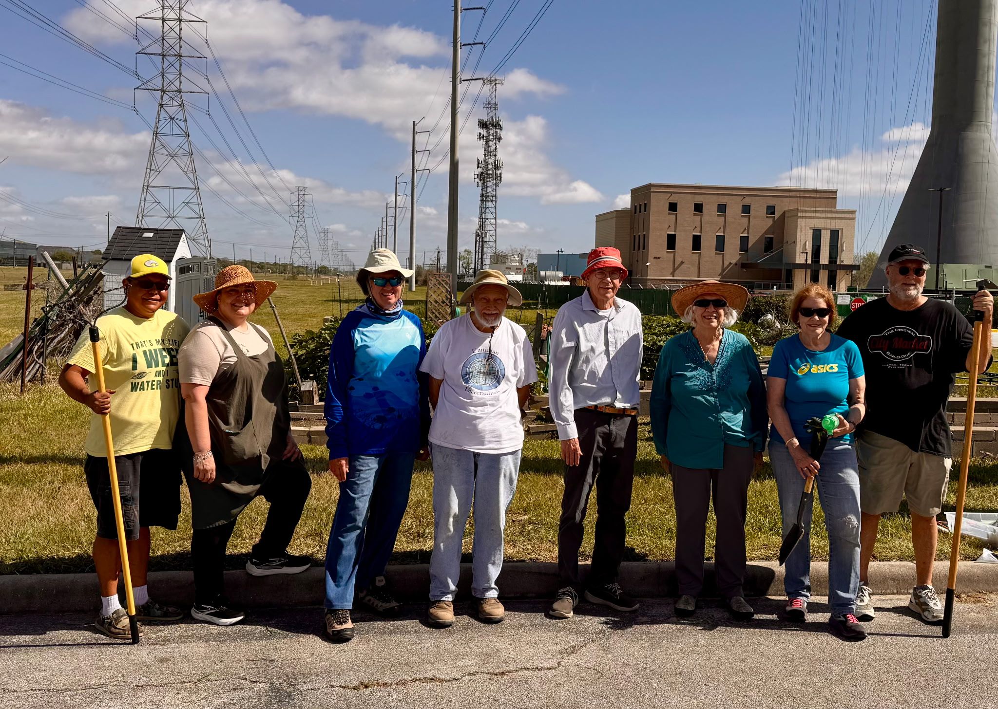 Group of nine people standing outdoors on a sunny day, some holding tools, with power lines and buildings in background.