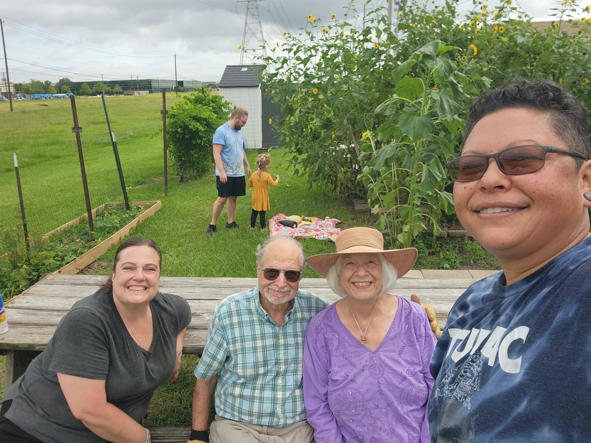 Four people sitting outdoors smiling, with a garden and a person with a child in the background, under a cloudy sky.
