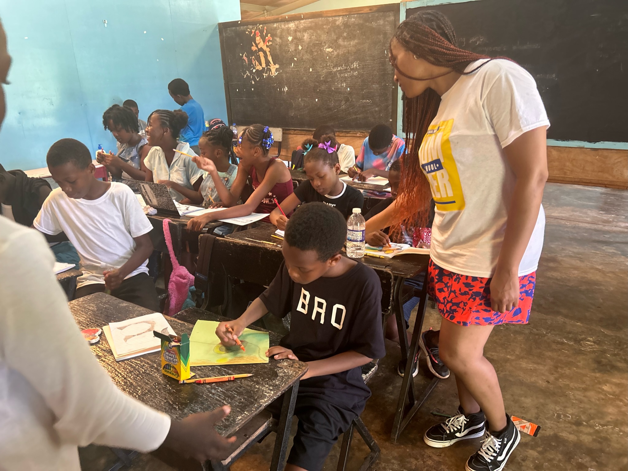 Teacher standing next to students seated at desks in classroom, blackboard in background, students writing or reading, diverse group, classroom setting
