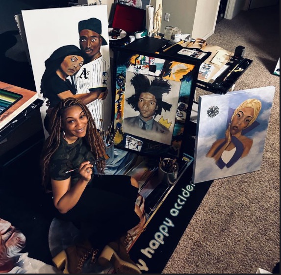 A woman with long dreadlocks sitting next to framed portraits on a table, with artwork and photos in the background.