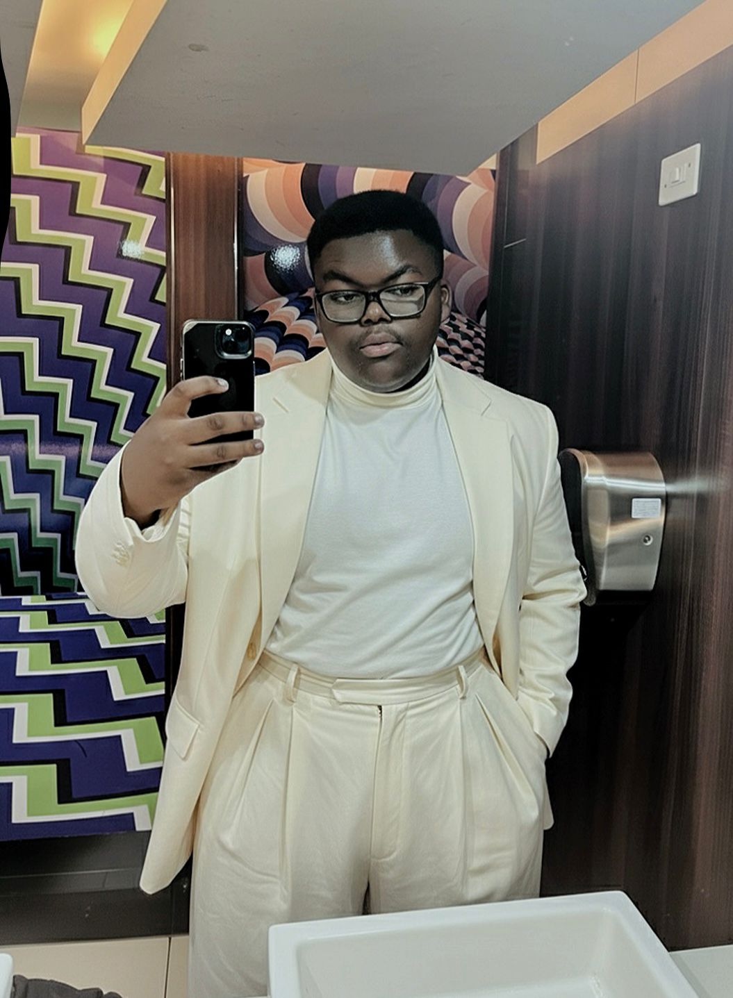 Person taking a mirror selfie in a room with colorful zigzag wall pattern, wearing a cream suit and glasses.