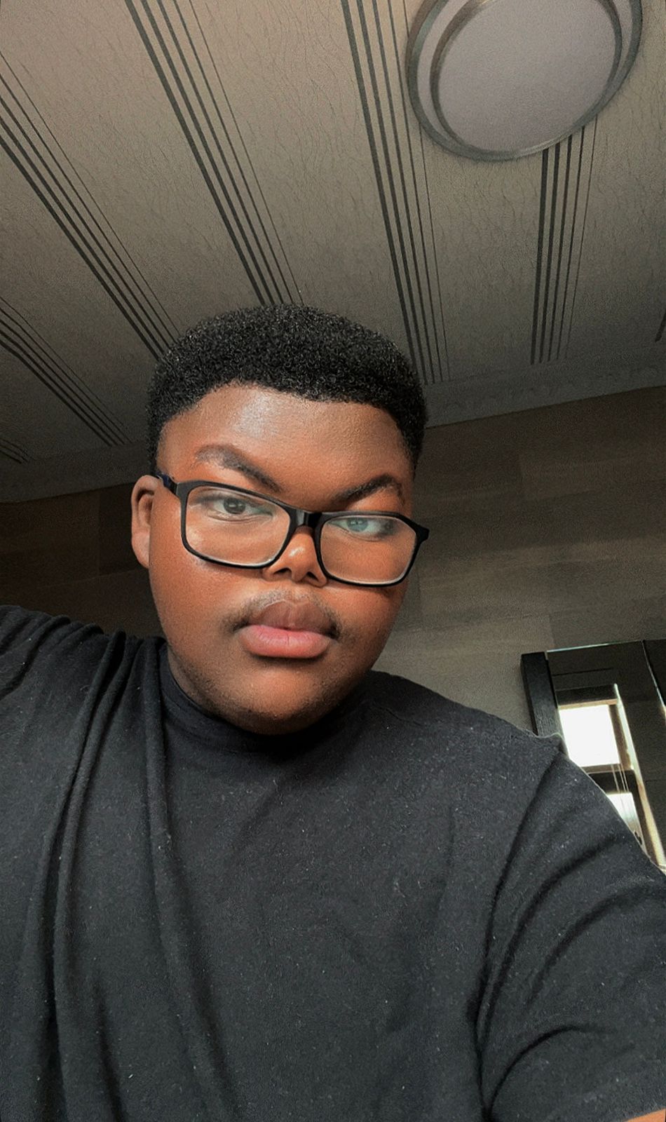 Young person with glasses and short curly hair wearing a black shirt, indoors with ceiling and window visible.