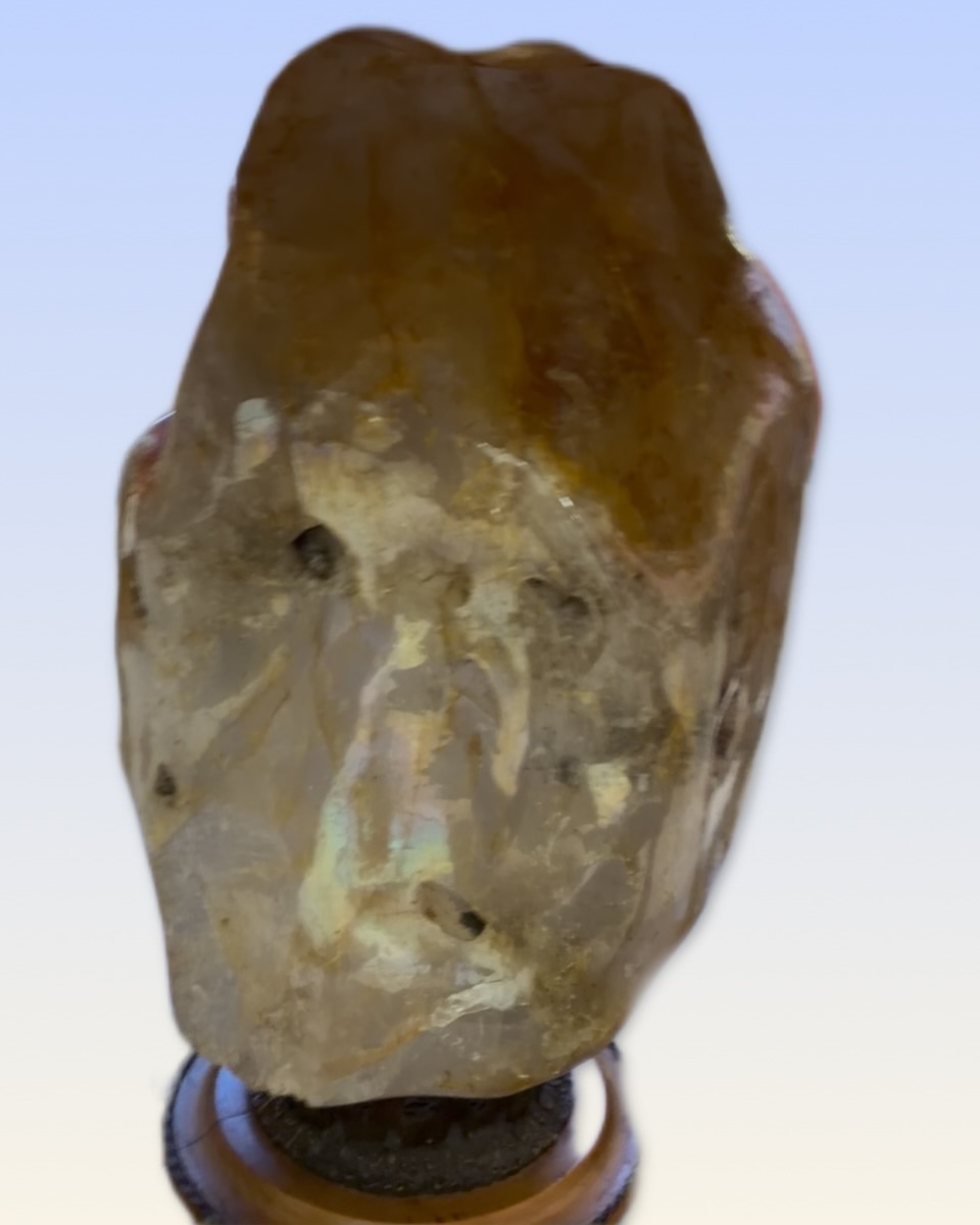A large, irregularly shaped stone sculpture with a face-like appearance, mounted on a base against a light blue background.