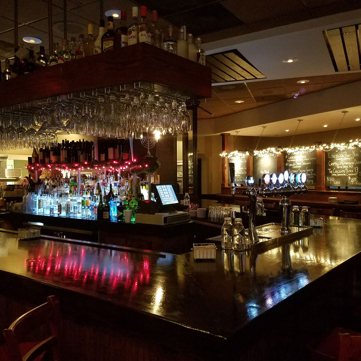 Bar with bottles, glasses, and a chalkboard menu, illuminated by warm lighting, with a reflective black countertop.