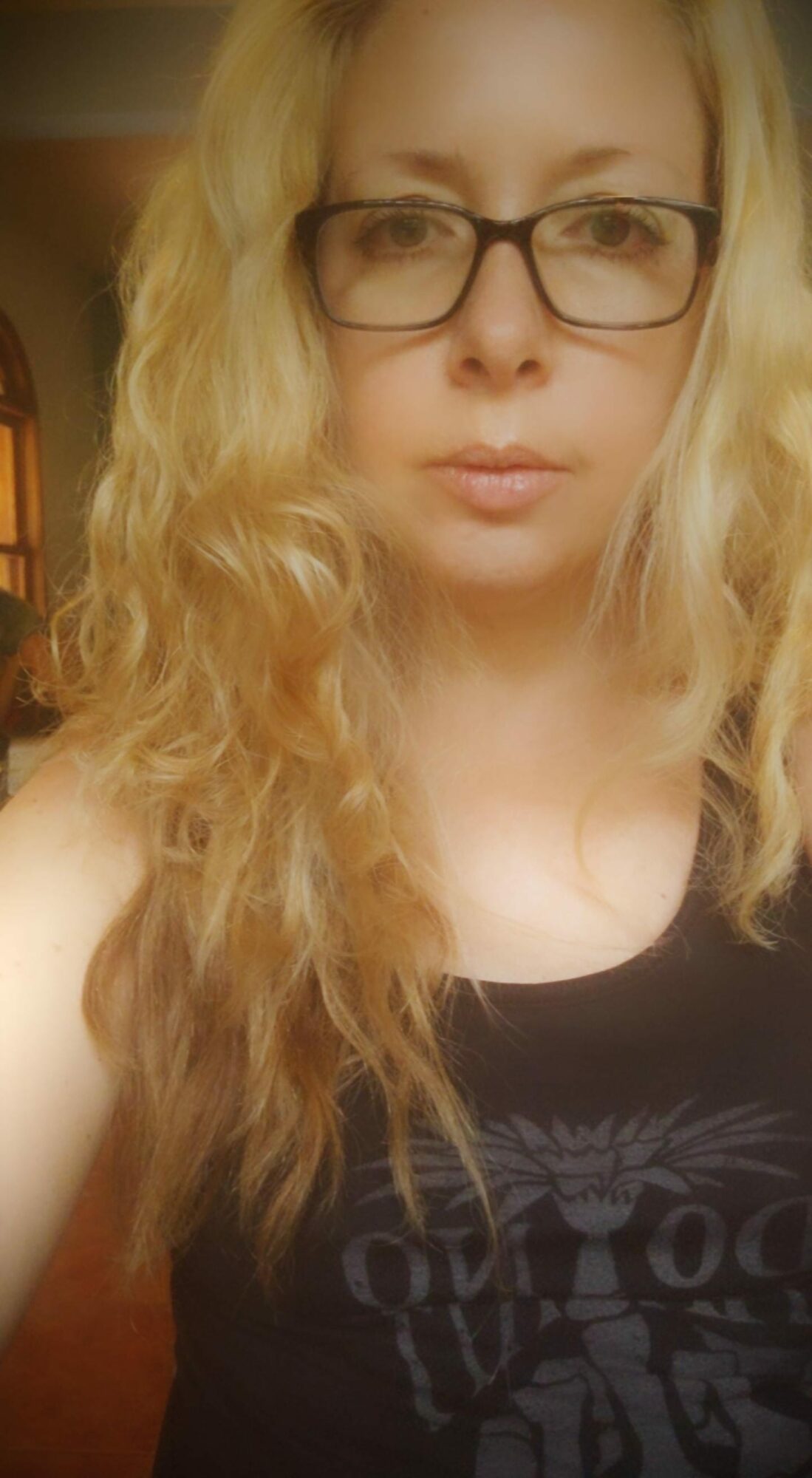 Woman with glasses and long wavy blonde hair, wearing a black top, indoors.