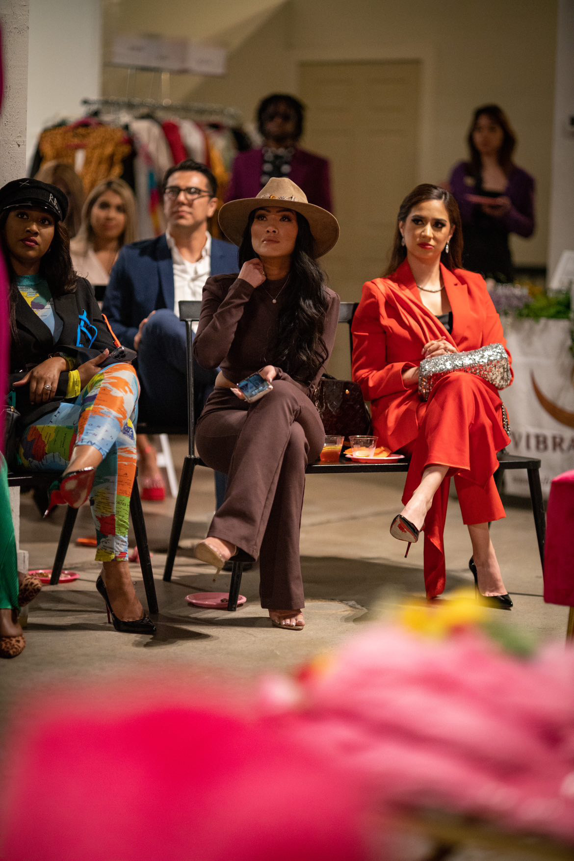 Group of women sitting and standing in a room, some wearing fashionable clothing and accessories, with clothing racks in the background.