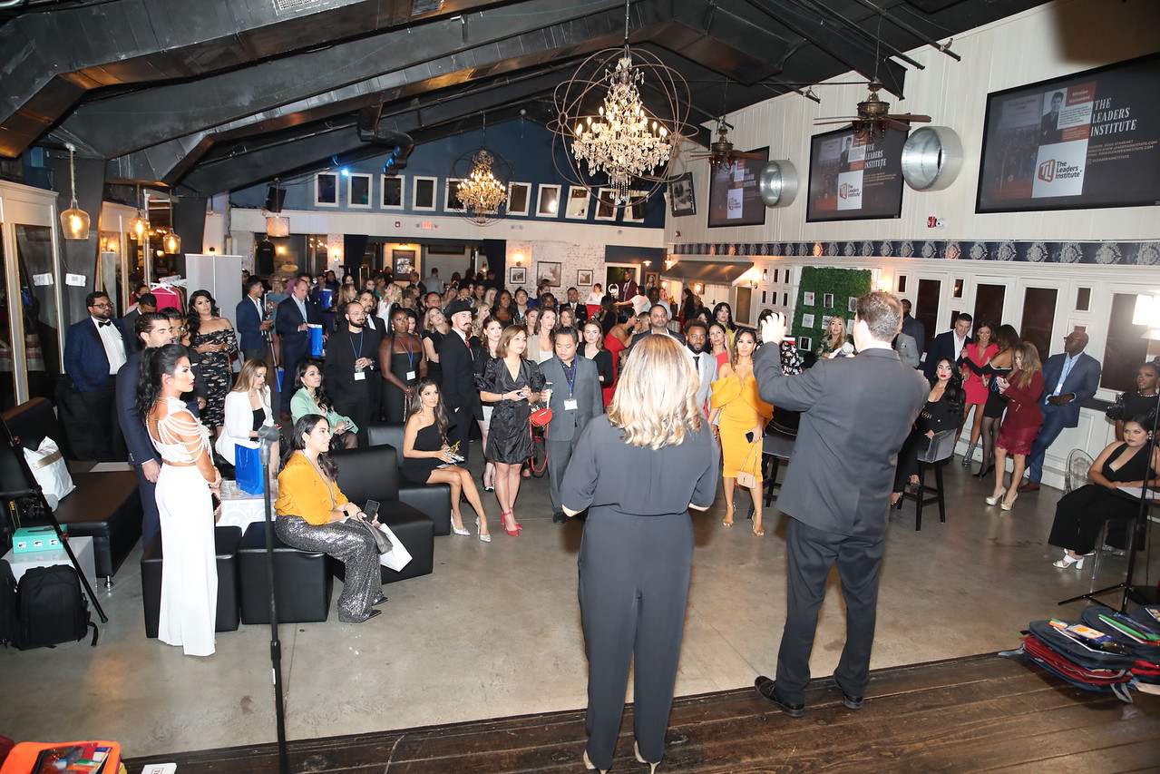 Crowd gathered in a decorated indoor venue with a chandelier, people sitting and standing, some taking photos, and screens on walls.