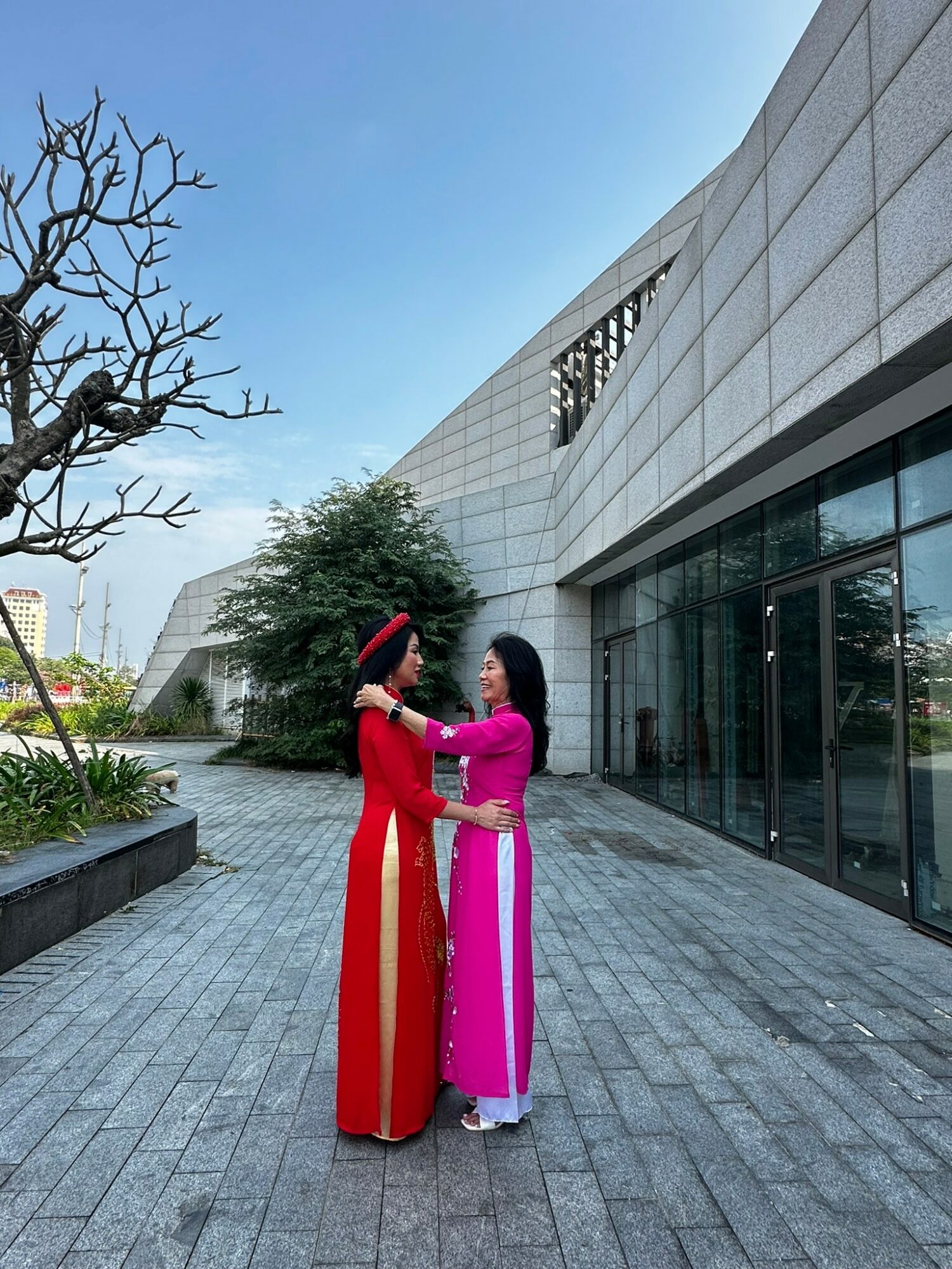 Two women in traditional dresses stand facing each other outdoors near a modern building, holding hands and smiling.