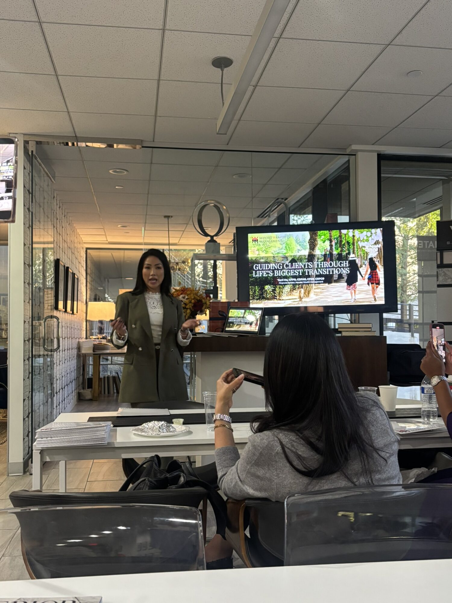 Woman standing and speaking in front of a group in a modern office or conference room, with a large screen displaying a presentation.