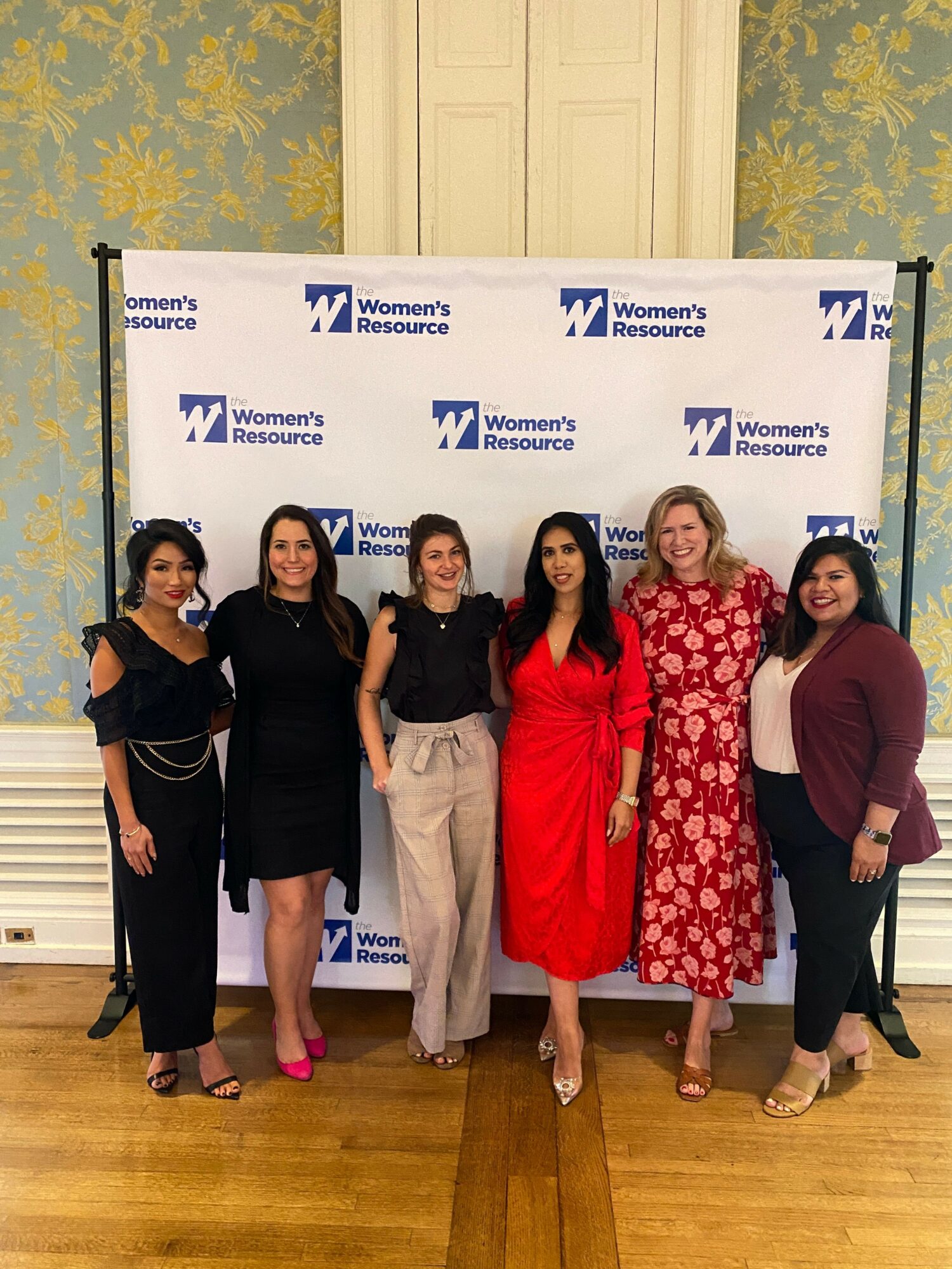 Six women standing in front of a backdrop with logos, dressed in formal and semi-formal attire, smiling at the camera.