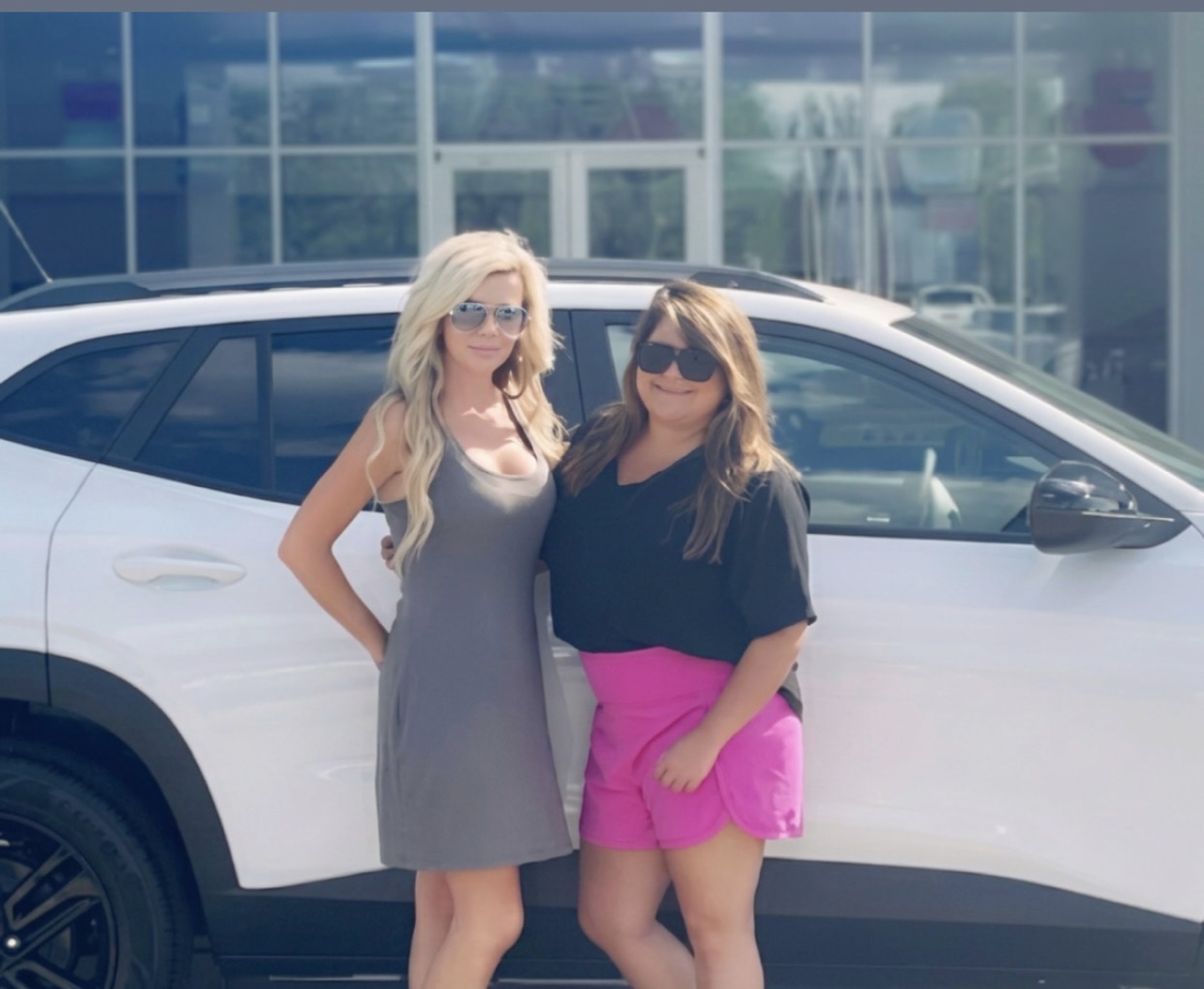 Two women stand in front of a white car, smiling, with a building and glass windows in the background.