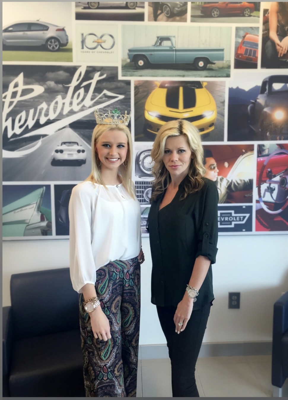 Two women standing in front of a collage of car images, smiling, one in a white blouse and patterned pants, the other in black attire.